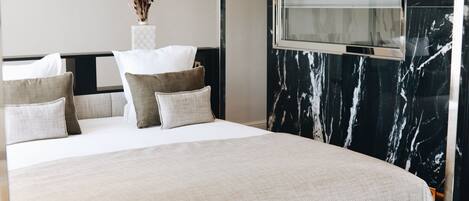 Una cama bien hecha con colcha blanca y beige, múltiples almohadas en tonos beige y blanco, y un florero con flores secas sobre una mesita de noche.