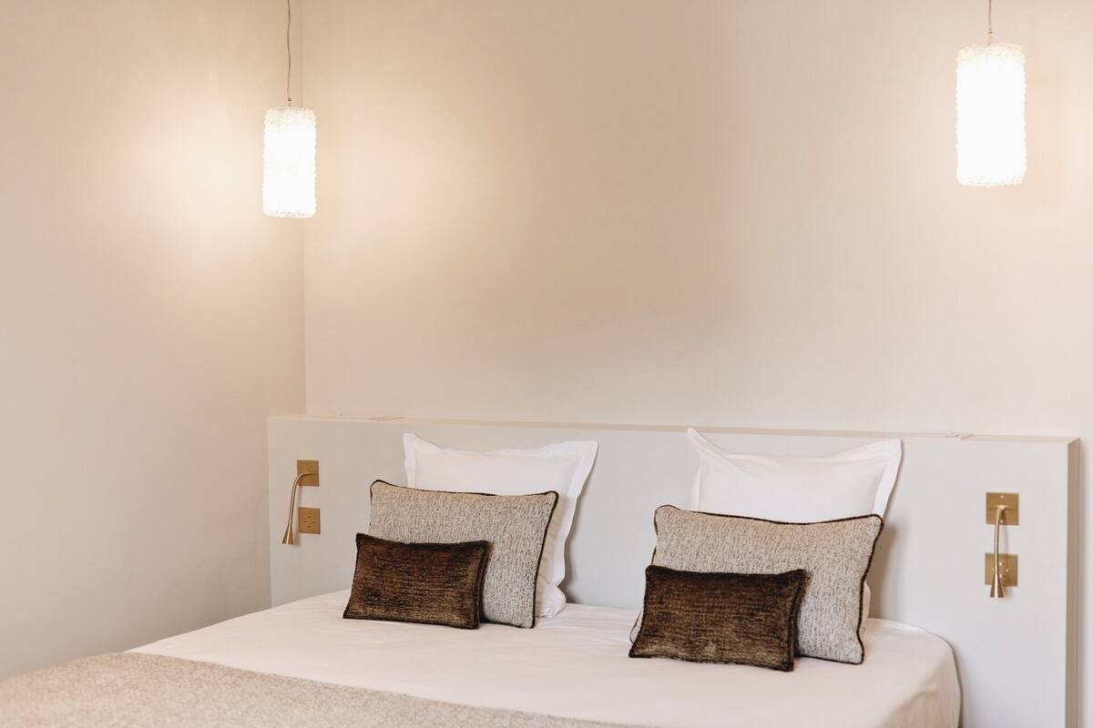 Una cama con ropa de cama blanca, dos almohadas con textura y dos lámparas colgantes.