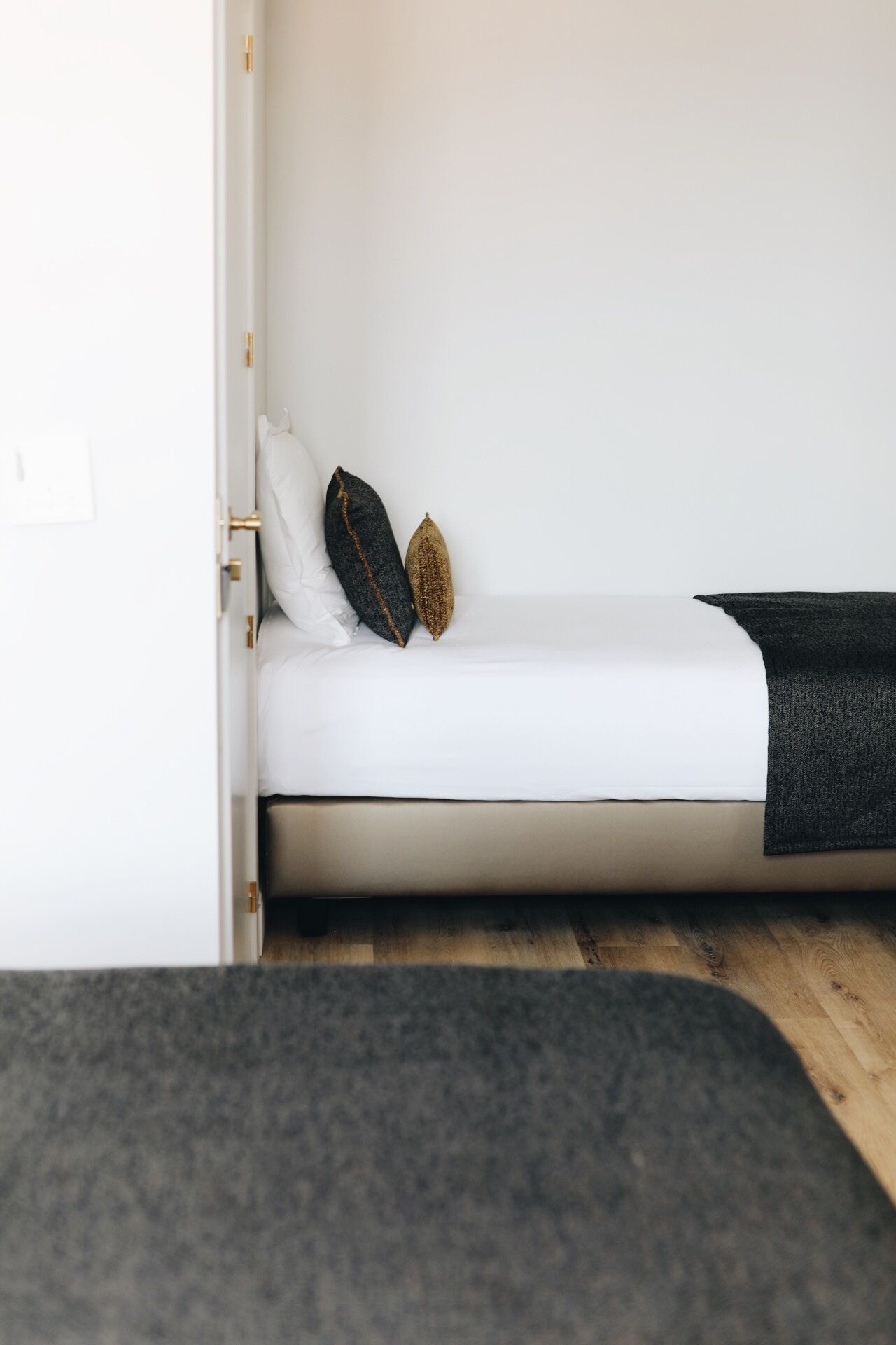 A small, compact bed with a white mattress and two pillows, one dark and one gold, in a minimalistic room with a wooden floor.
