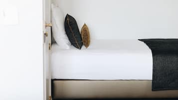 Een klein, compact bed met een witte matras en twee kussens, één donker en één goud, in een minimalistische kamer met een houten vloer.