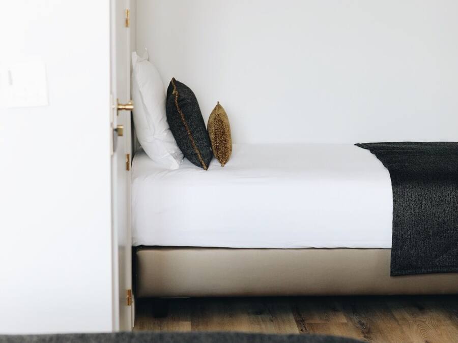 Een klein, compact bed met een witte matras en twee kussens, één donker en één goud, in een minimalistische kamer met een houten vloer.