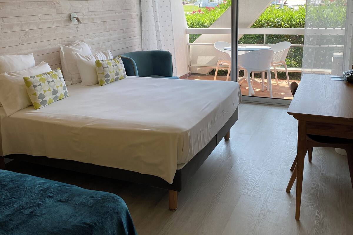 A hotel room with a large bed, a smaller bed, a desk, and a balcony with a view of greenery.