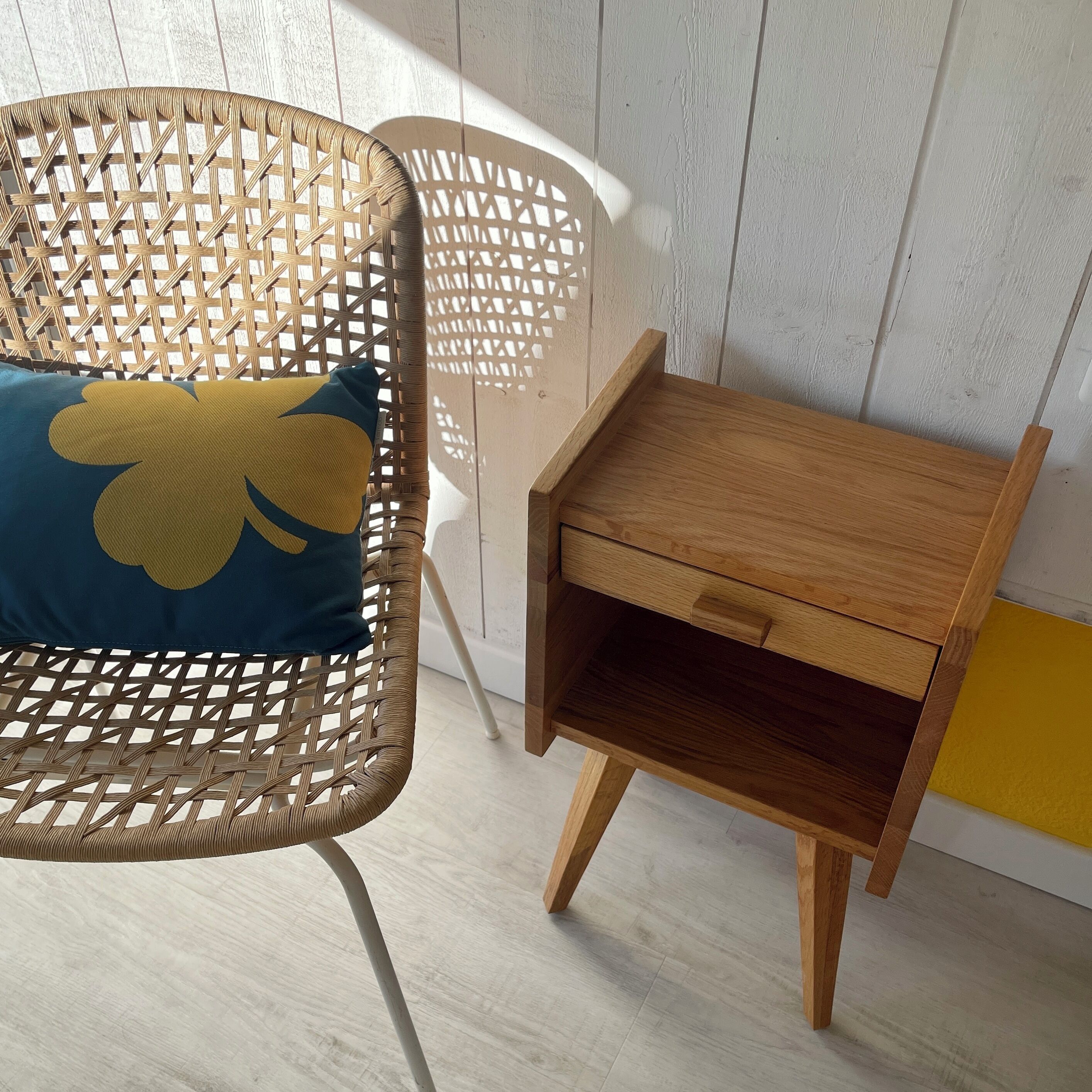 Een rieten stoel met een kussen en een houten nachtkastje met een lade.