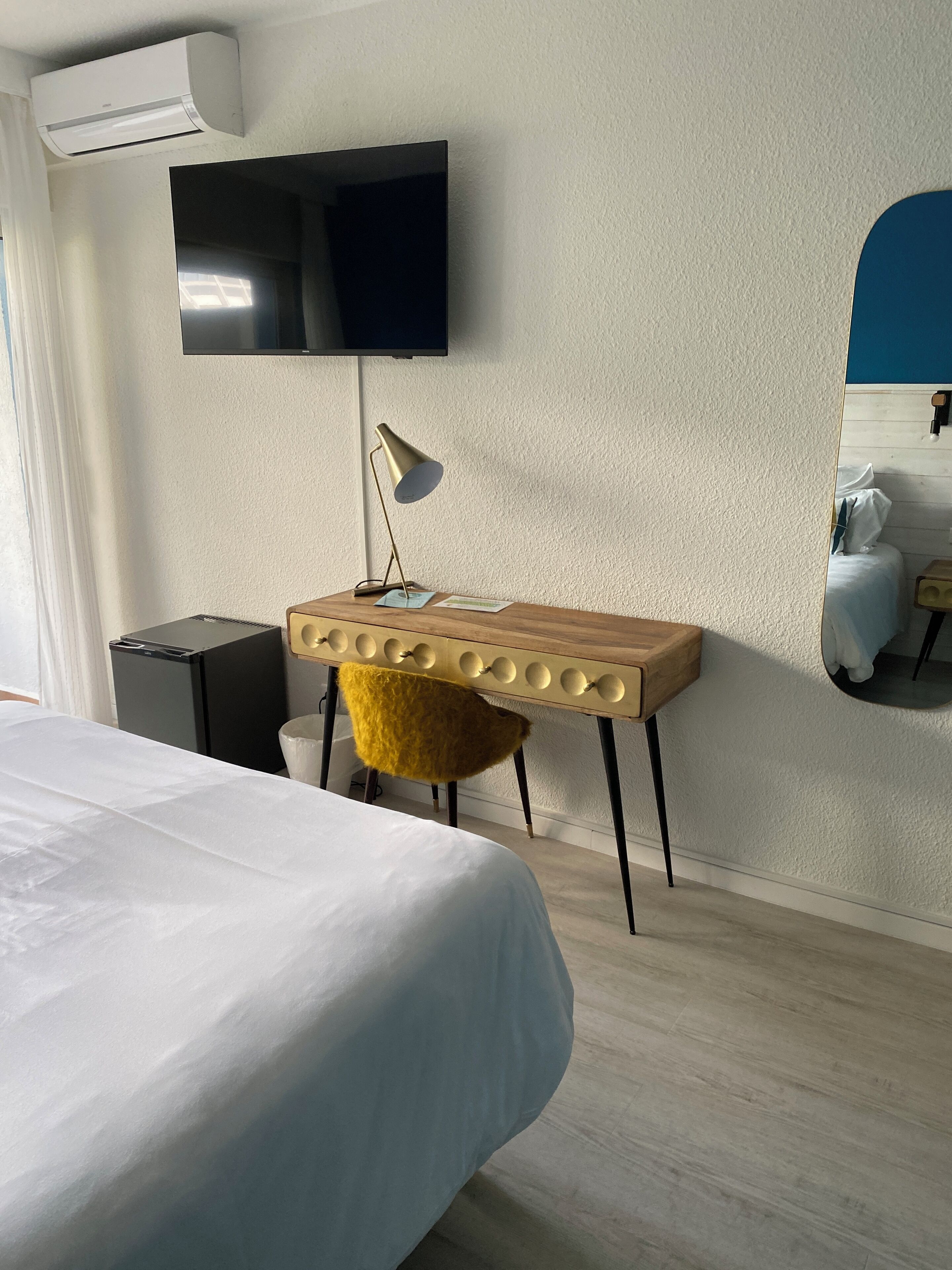 A hotel room with a bed, a desk with a lamp, a chair, a television mounted on the wall, and a mirror.