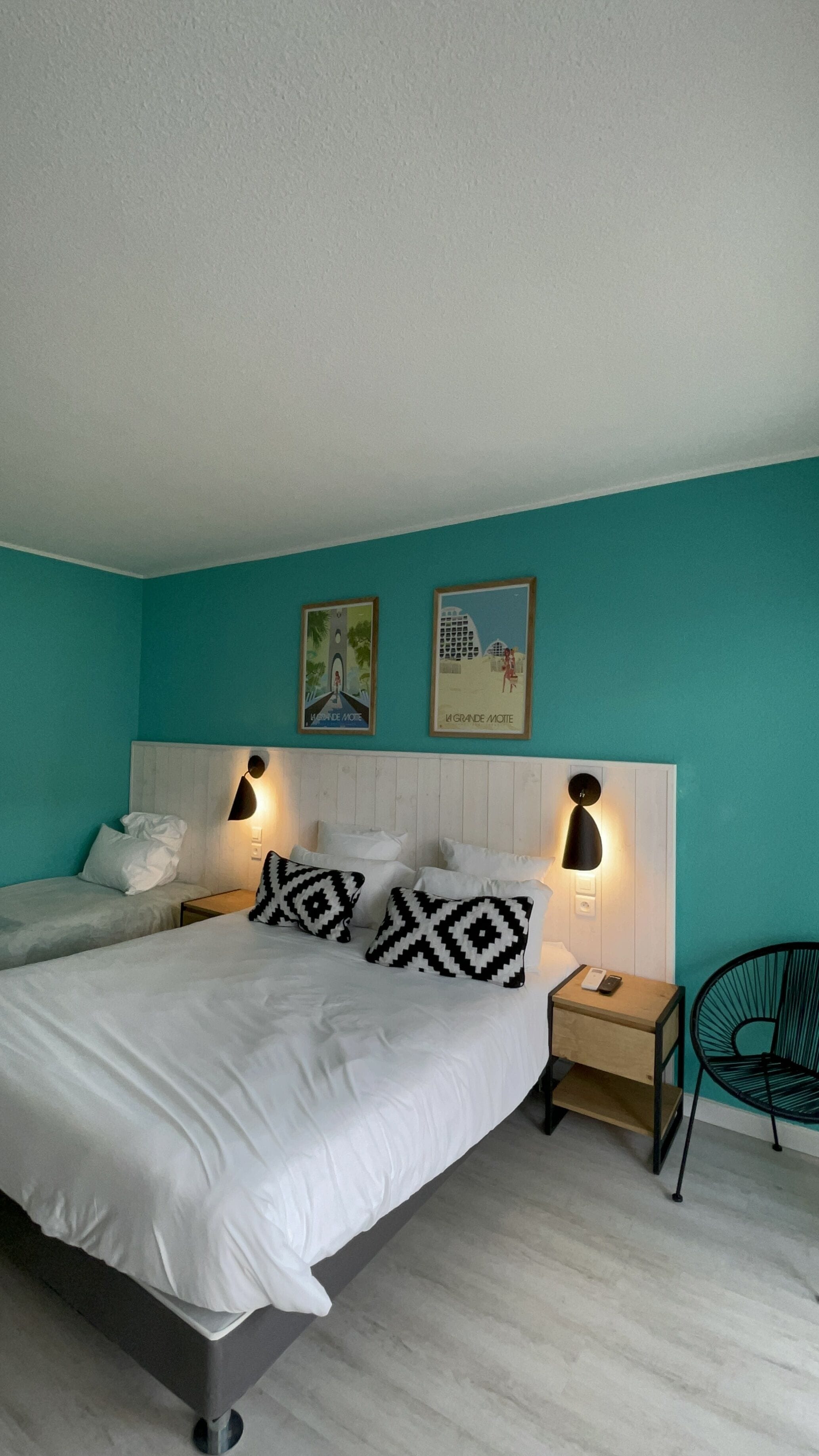 A bedroom with two beds, teal walls, and a white headboard. There are two wall-mounted lamps and a framed picture above the beds.