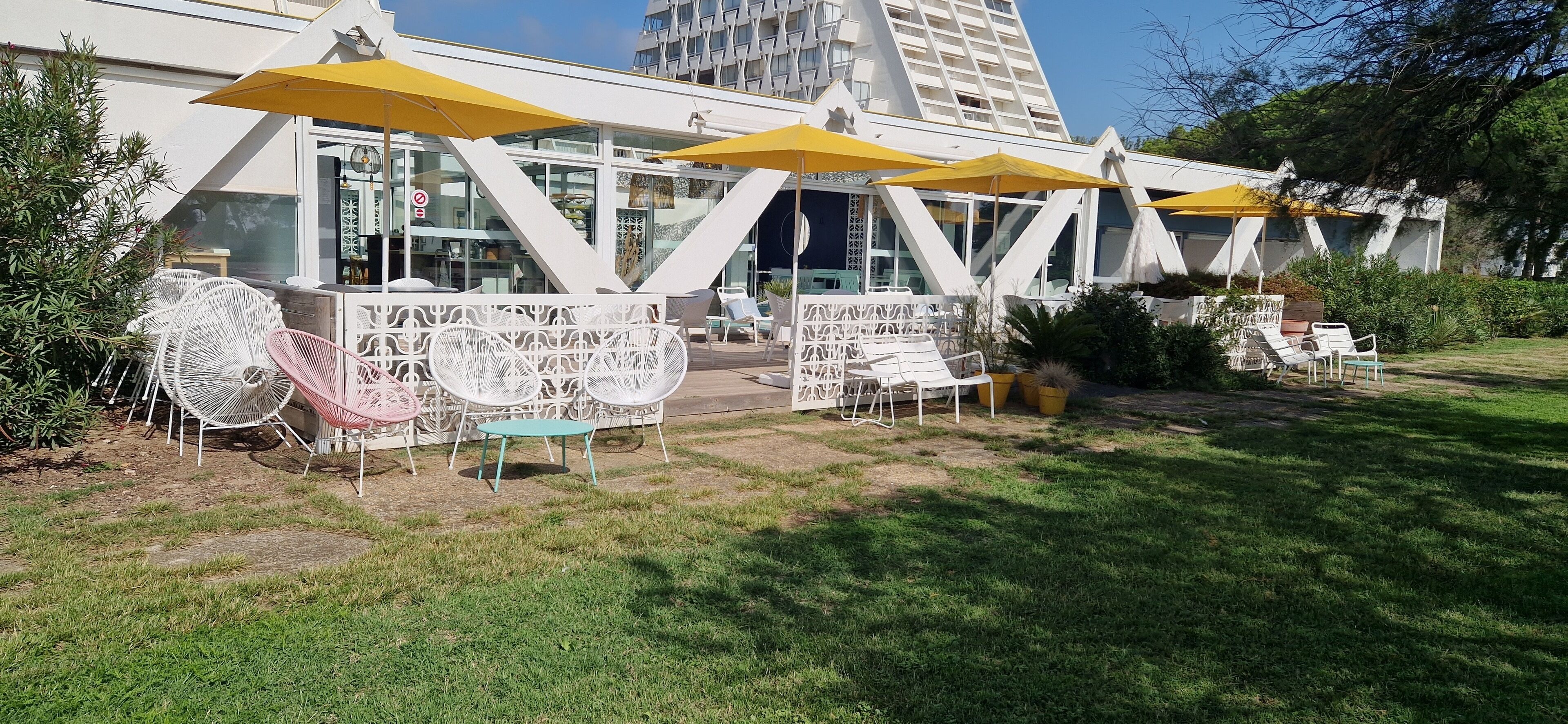An outdoor seating area with white chairs and yellow umbrellas, a glass building in the background, and a grassy lawn.