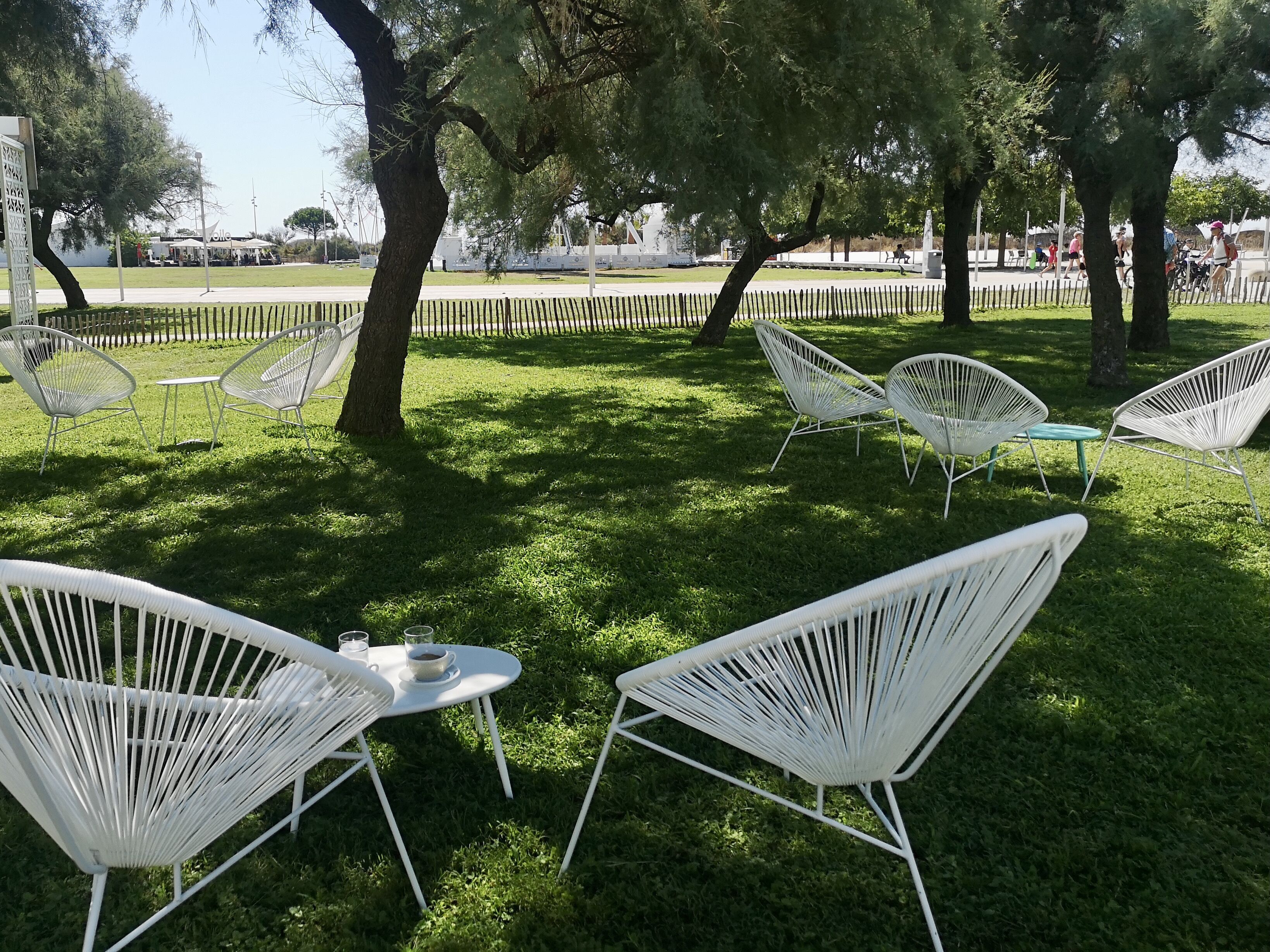 Un área de estar al aire libre con sillas y mesas de metal blanco, rodeada de césped y árboles.