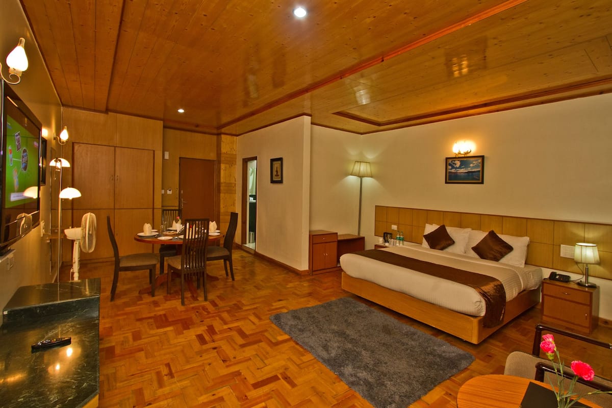 A hotel room with a wooden ceiling, a bed with a headboard, a bedside table with a lamp, a small table with chairs, and a dining area with a table and chairs.