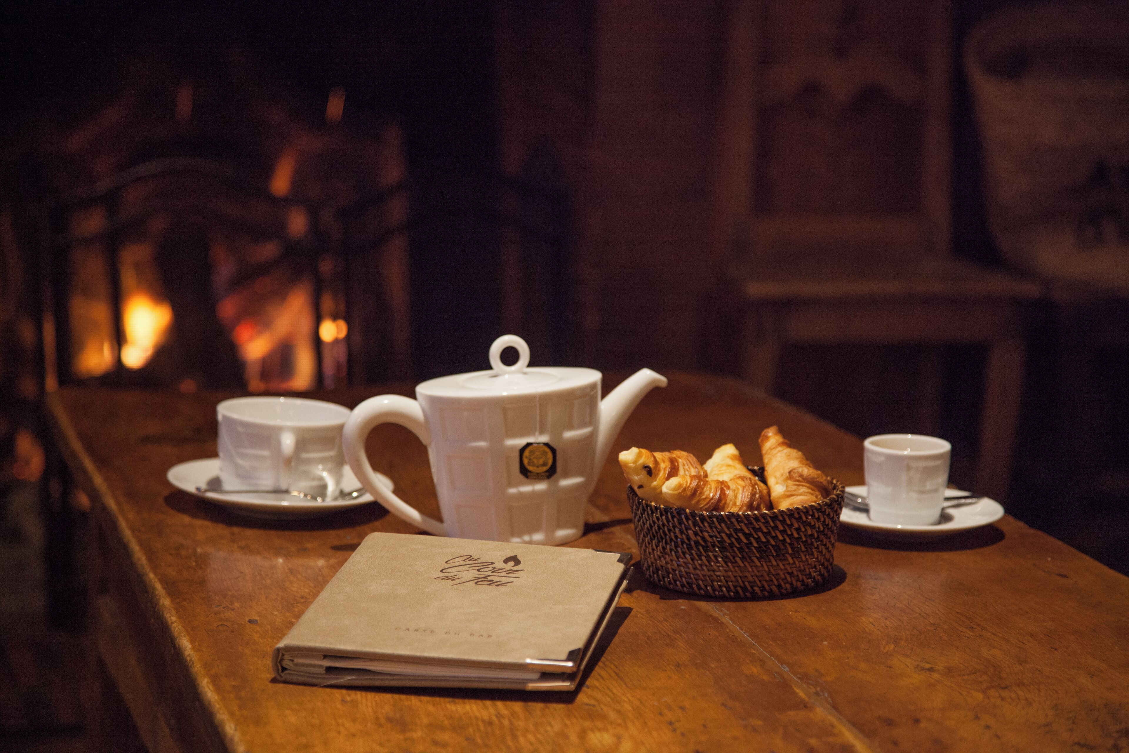 A wooden table with a teapot, two cups, a basket of croissants, and a menu.