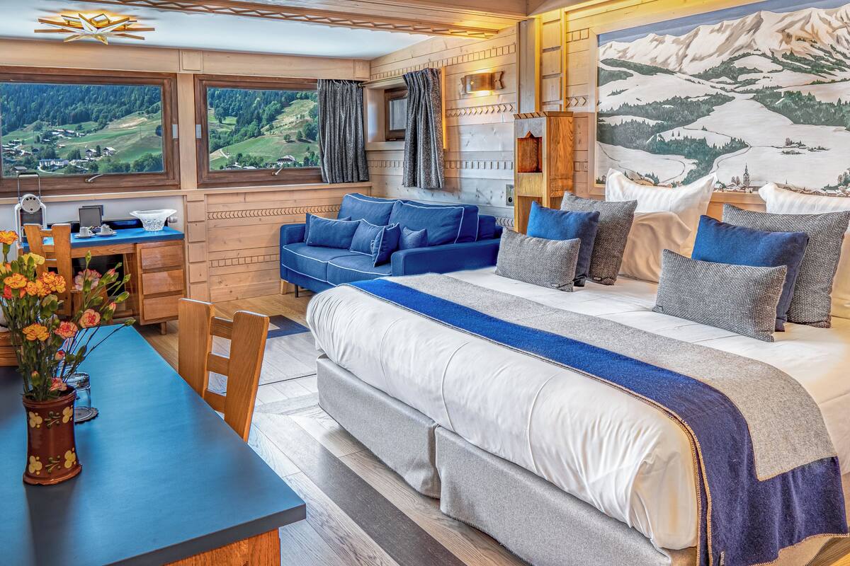 Ein gemütliches Zimmer mit einem großen Bett, einem blauen Sofa, einem Holztisch und Blick auf eine gebirgige Landschaft durch die Fenster.