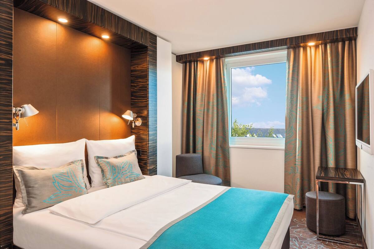 A hotel room with a large bed, a TV, a window with curtains, and a view of the outdoors.