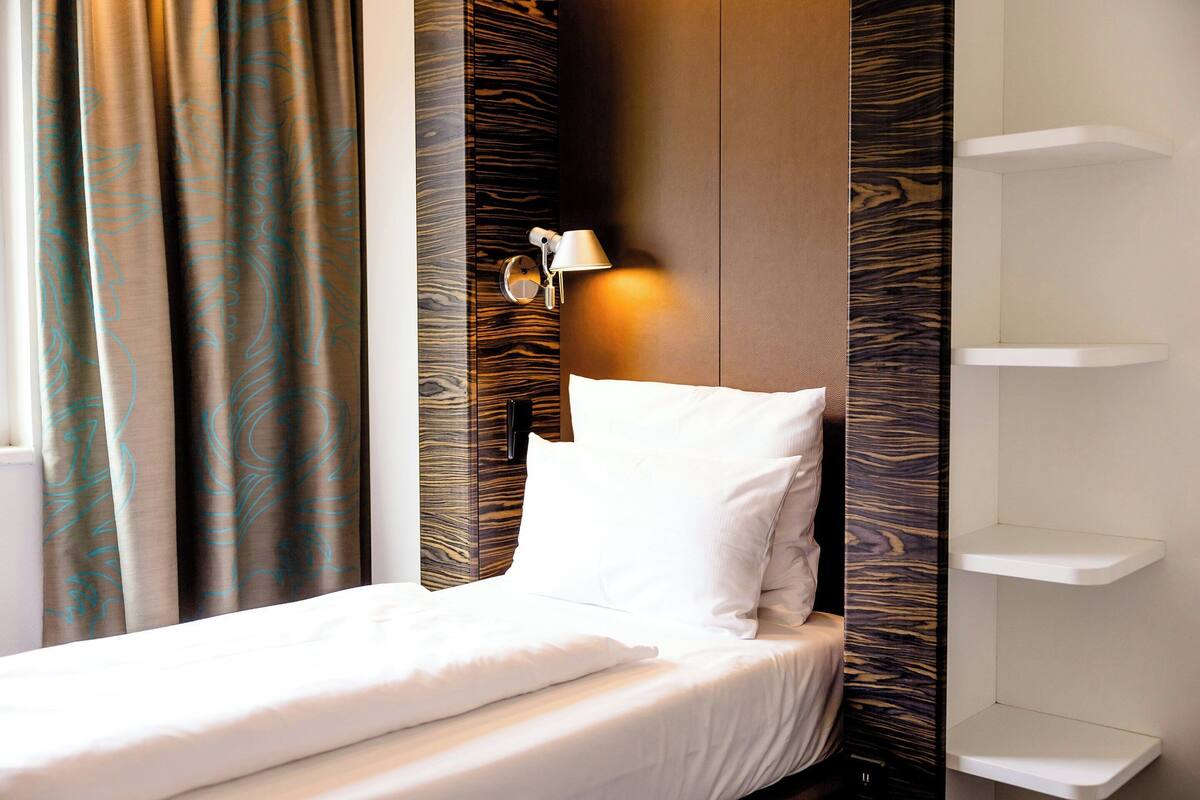 A hotel room with a bed, a nightstand, a wall-mounted lamp, and a window with curtains.