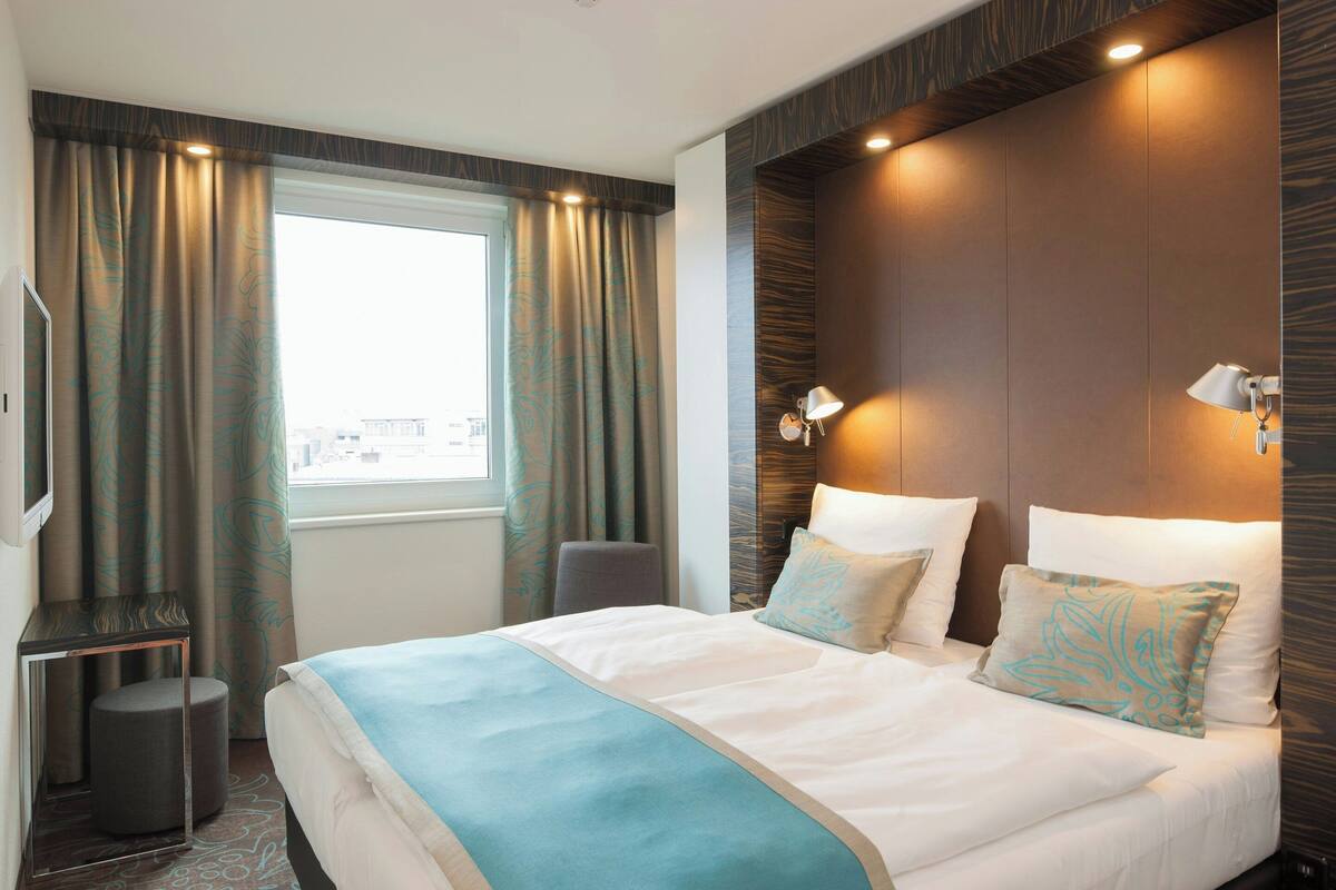 A hotel room with a large bed, a desk, a chair, and a view of the city through the window.