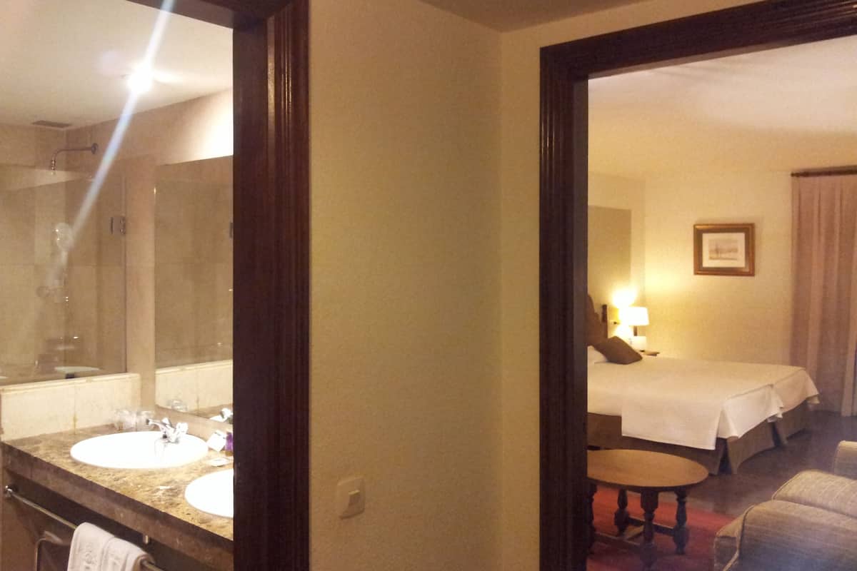 Une chambre d’hôtel avec un grand miroir, une salle de bain avec un lavabo et un porte-serviettes, un lit recouvert d’une courtepointe blanche, une petite table ronde et une chaise.