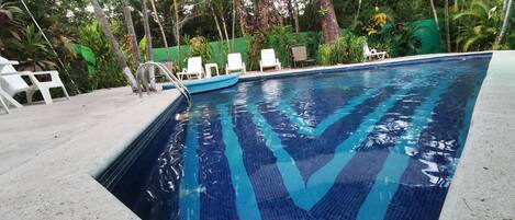 A swimming pool with clear blue water, surrounded by white lounge chairs and greenery.