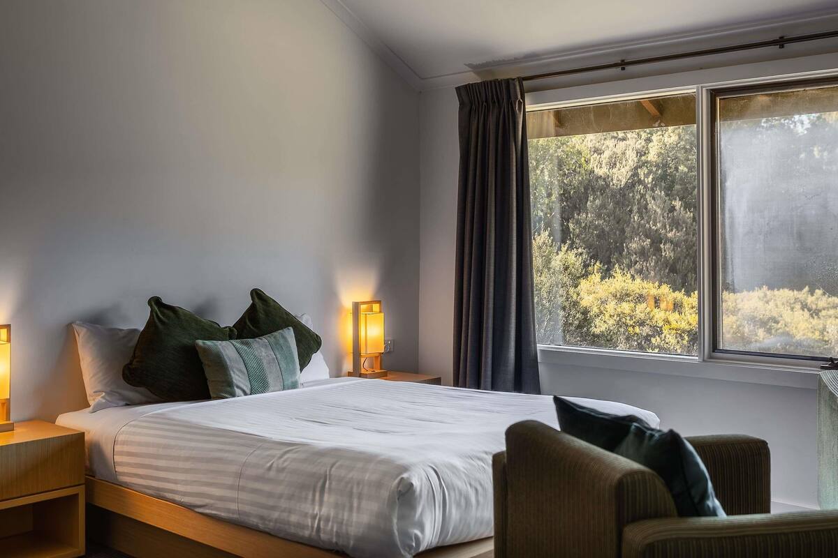 Een slaapkamer met een bed, nachtkastjes, een stoel, een raam met gordijnen en uitzicht op bomen.