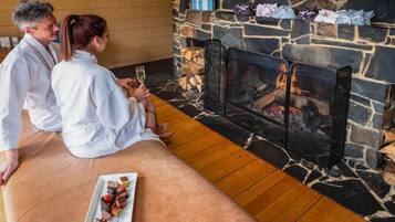 Un couple est assis près d’une cheminée en pierre, avec un plateau rempli de chocolats et de fraises.