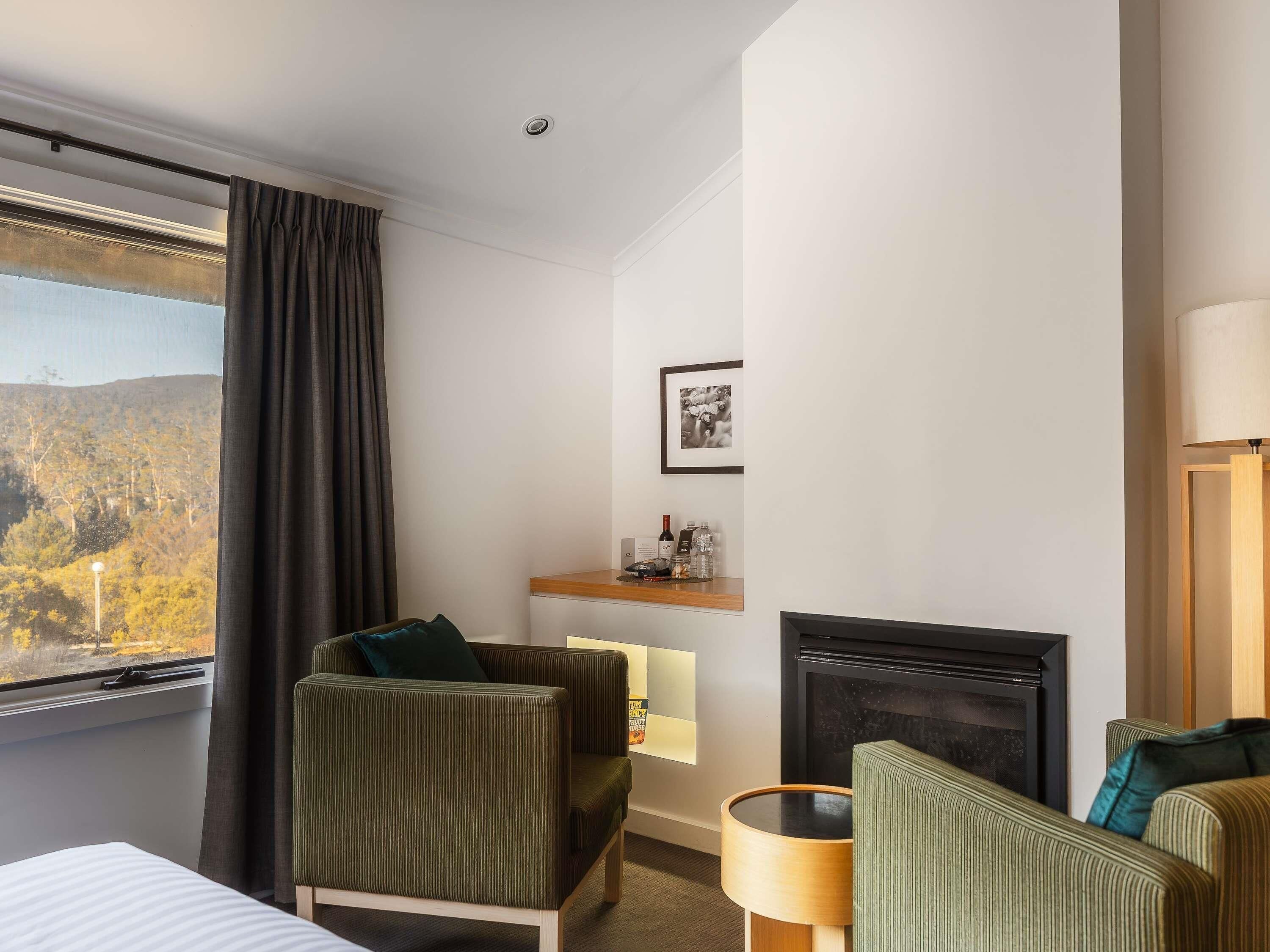A hotel room with a bed, two armchairs, a small table, a fireplace, a window with curtains, and a picture on the wall.