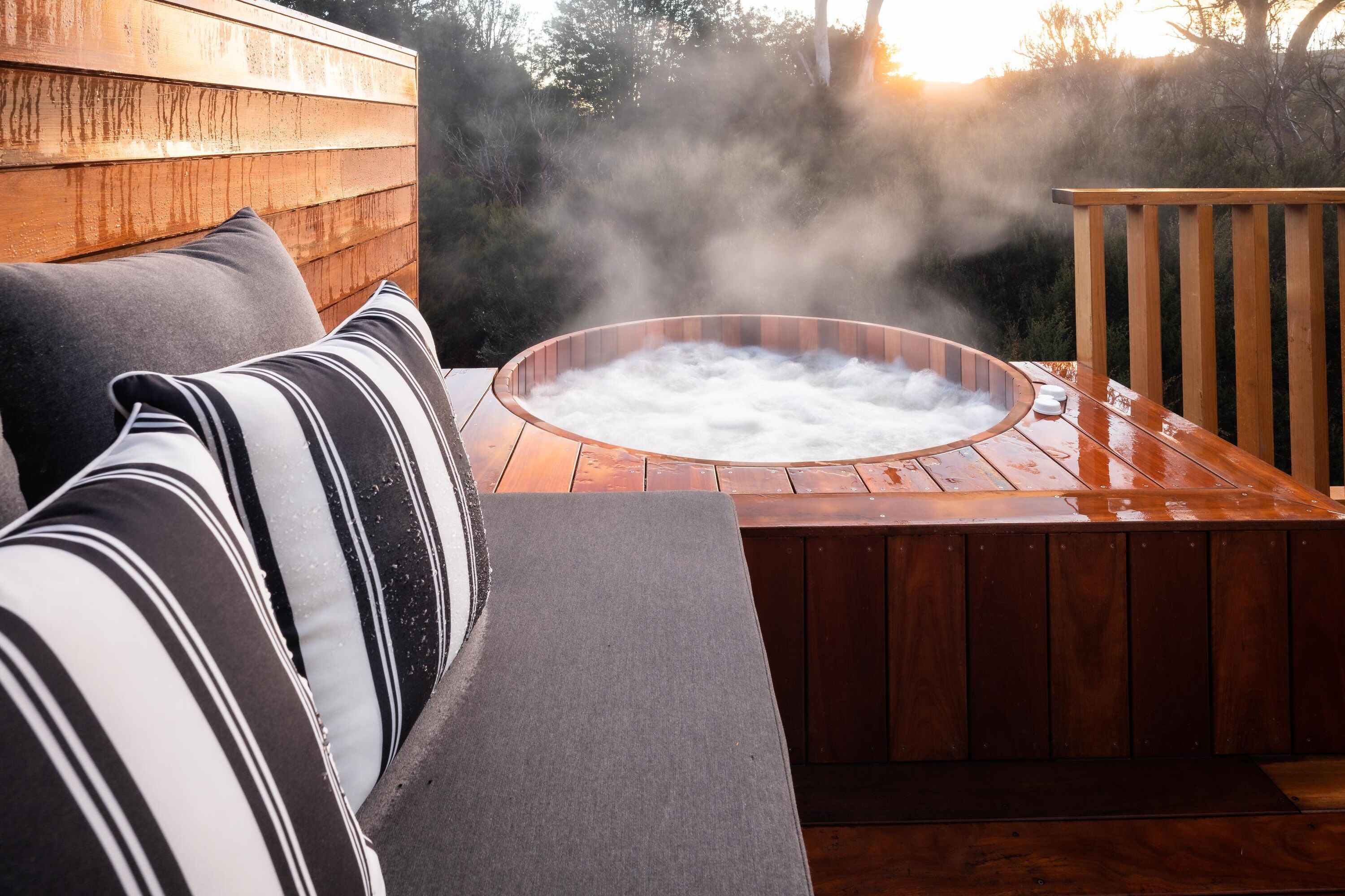 A hot tub with steam rising, surrounded by wooden deck and a sofa with striped cushions.