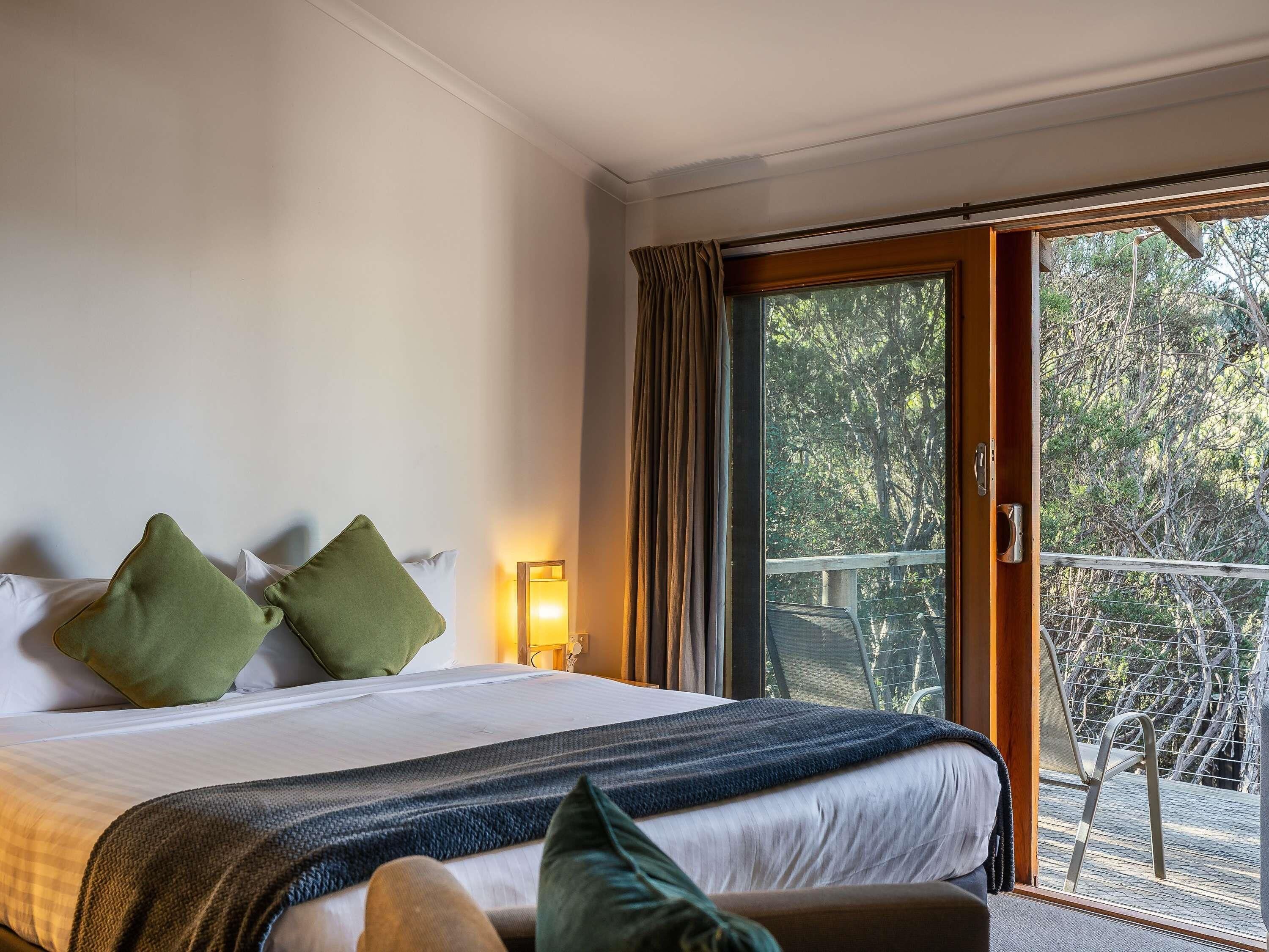 Une chambre d’hôtel avec un lit, une table de chevet, une lampe et une vue sur un balcon bordé d’arbres.