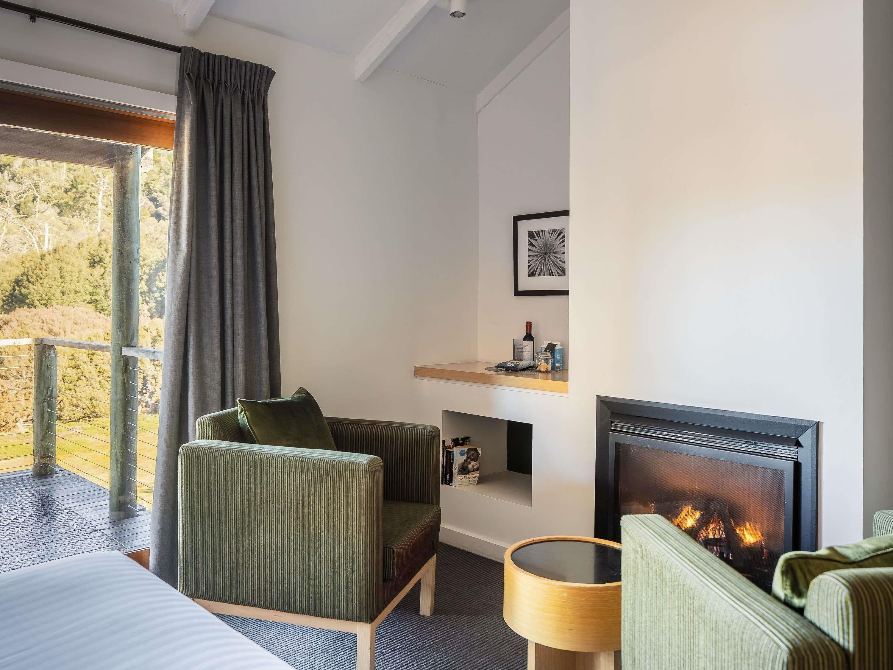 A hotel room with a bed, two green upholstered chairs, a fireplace, a window with curtains, and a shelf with items.