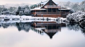 Una cabaña de madera con un balcón grande, ubicada junto a un lago tranquilo y árboles nevados al fondo.