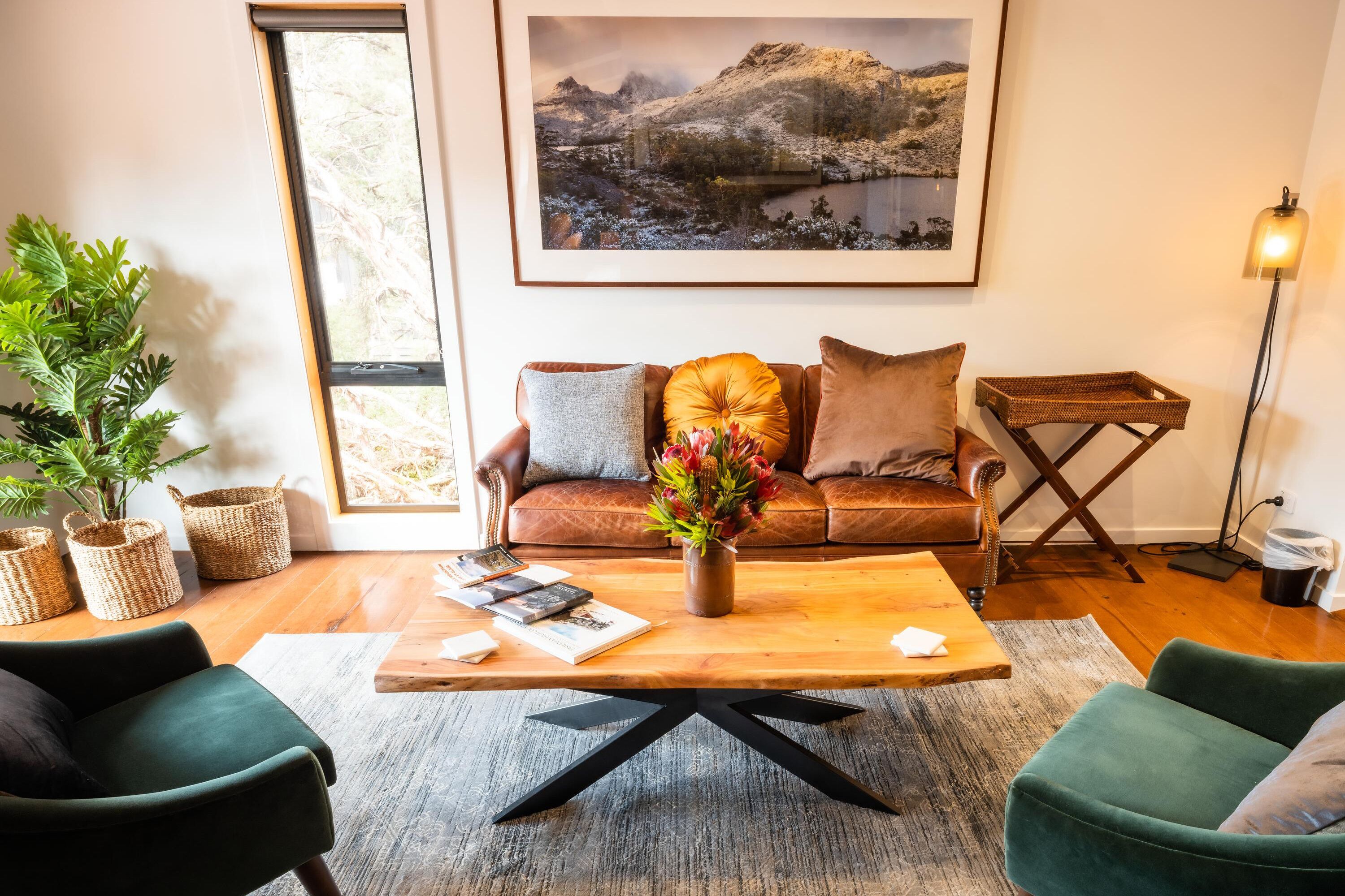 A cozy living room with a brown leather sofa, green armchairs, a wooden coffee table, and a large framed landscape painting.
