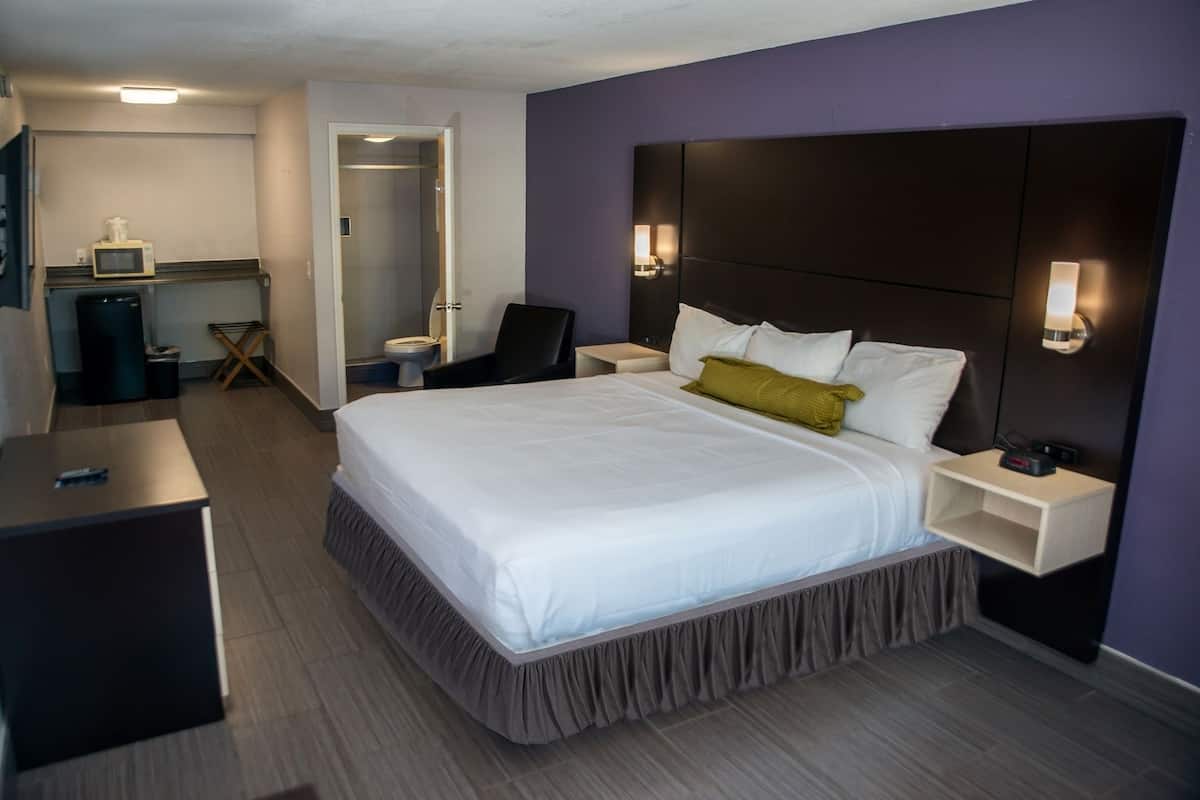 A hotel room with a large bed, bedside tables, a desk, a chair, a TV, and a bathroom visible through an open door.