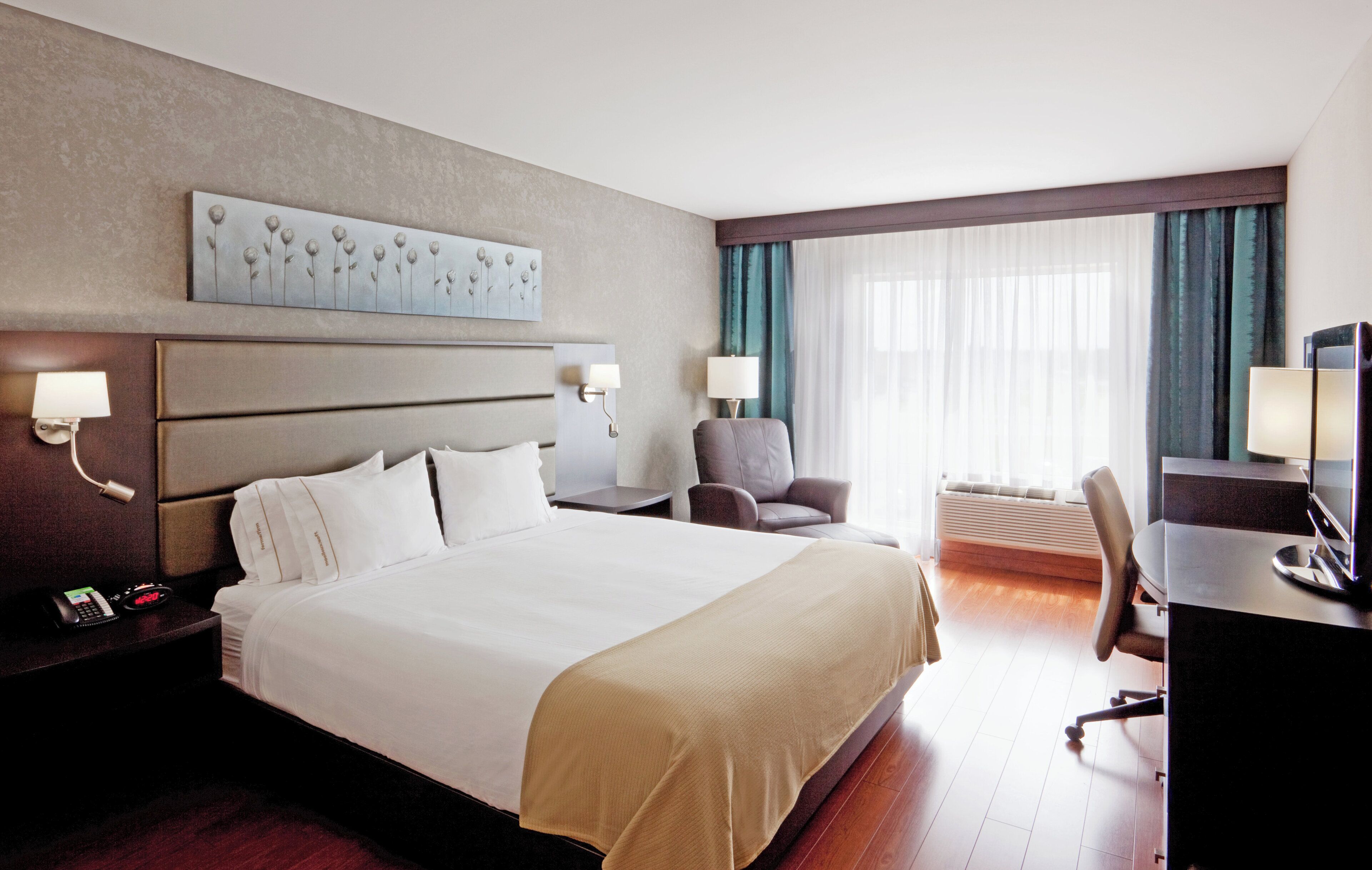 A hotel room with a large bed, bedside tables, a desk with a computer, a chair, and a window with curtains.