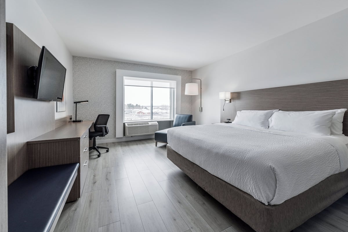 A modern hotel room with a large bed, a desk with a chair, a flat-screen TV mounted on the wall, and a large window offering a view of the city.