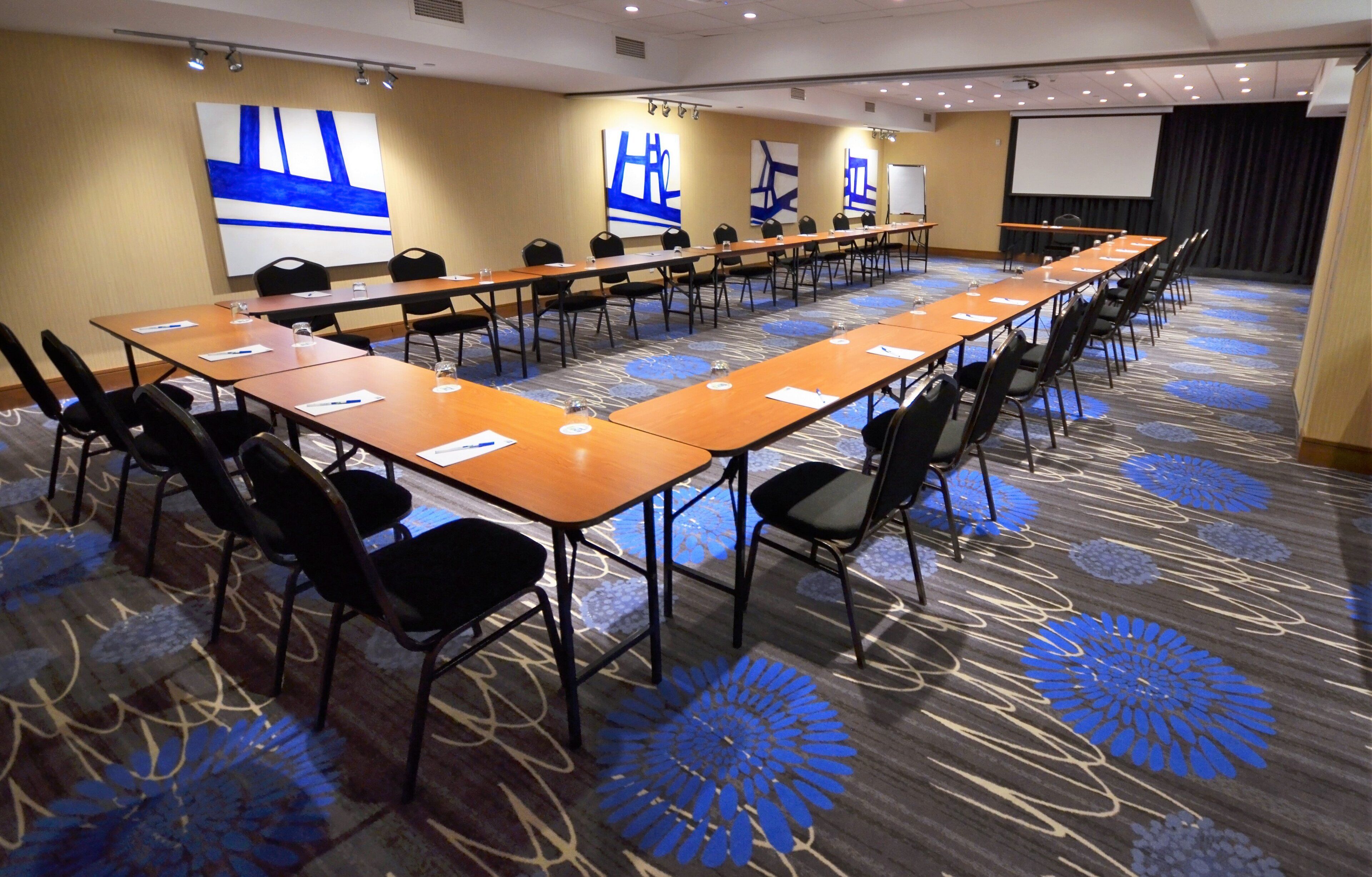 A conference room with long tables and chairs, a projector screen, and abstract wall art.