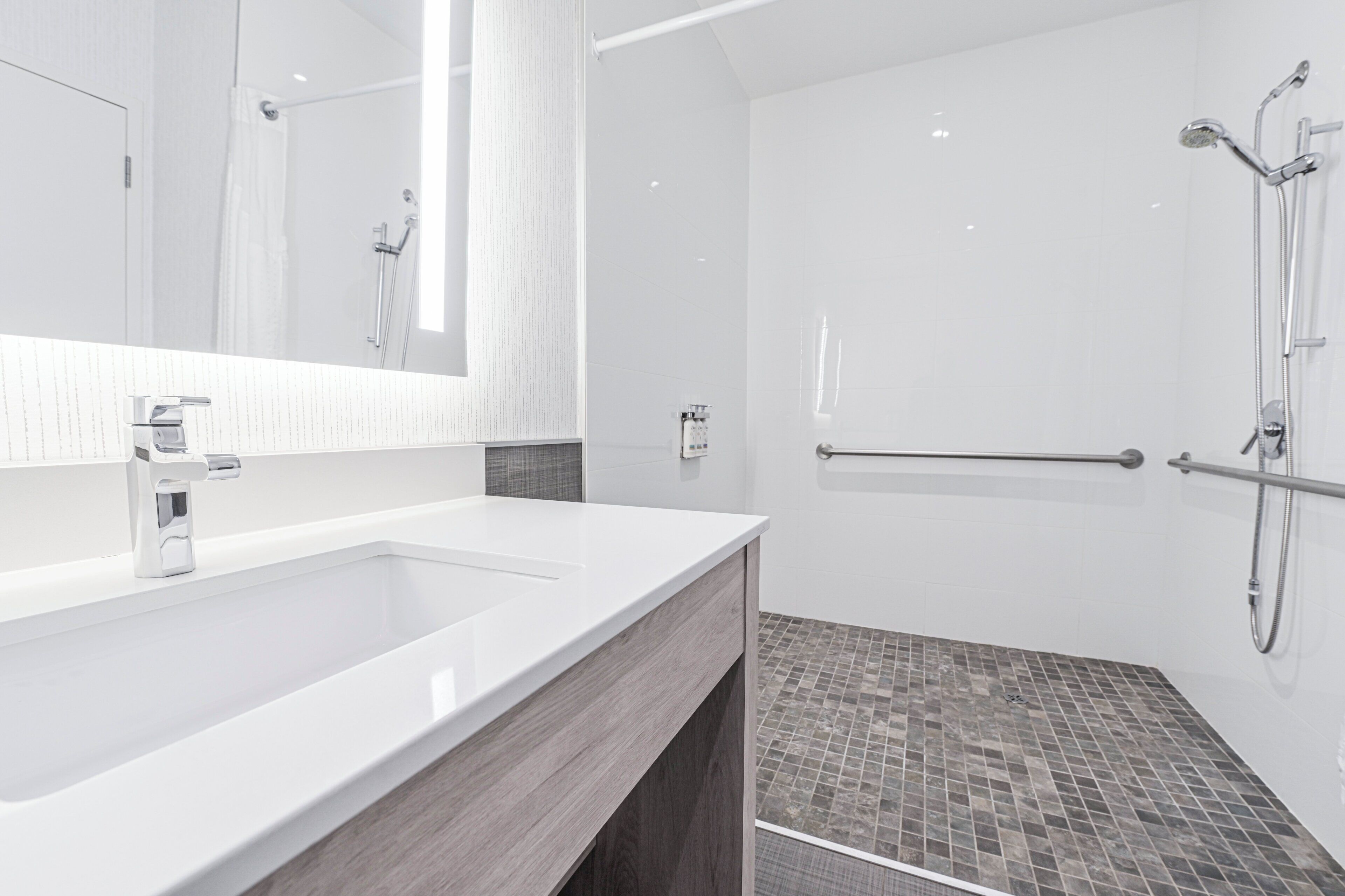 Une salle de bain moderne avec un grand miroir, un lavabo blanc et un espace douche équipé d’une barre d’appui.
