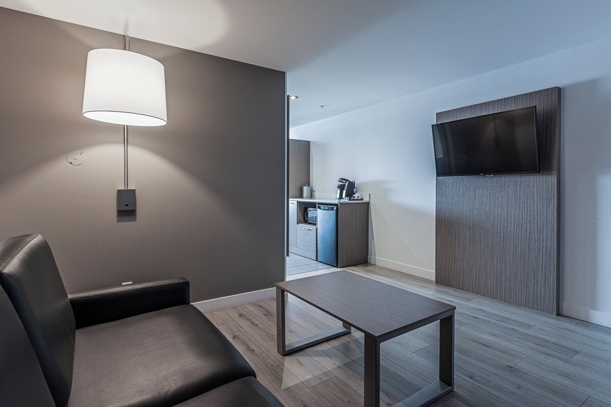 Ein modernes Hotelzimmer mit einer Couch, einem Tisch, einem an der Wand befestigten Fernseher und einer Küchenzeile im Hintergrund.