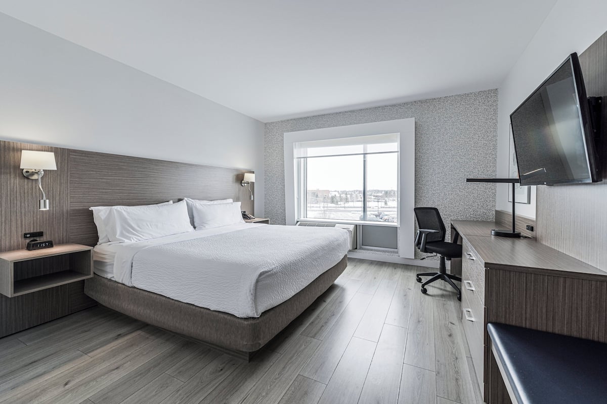 A modern hotel room with a large bed, a desk, a TV, and a window with a city view.