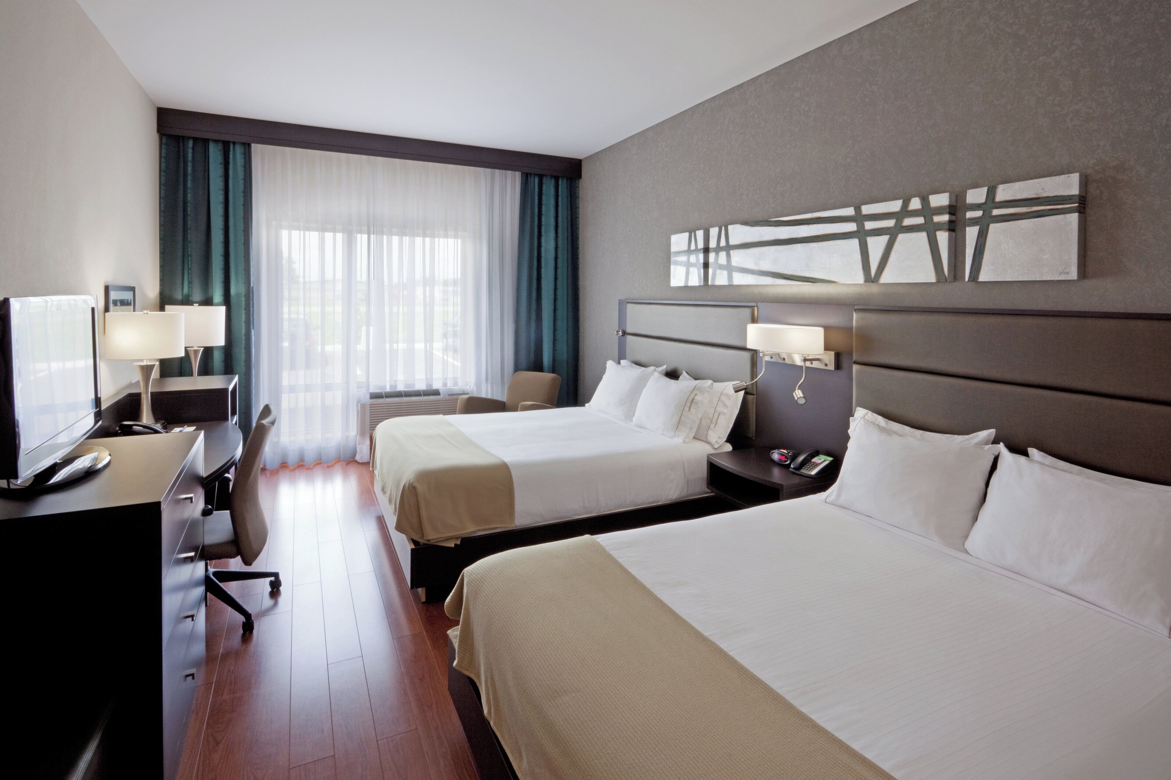 A hotel room with two beds, a desk with a lamp, a chair, and a large window with curtains.