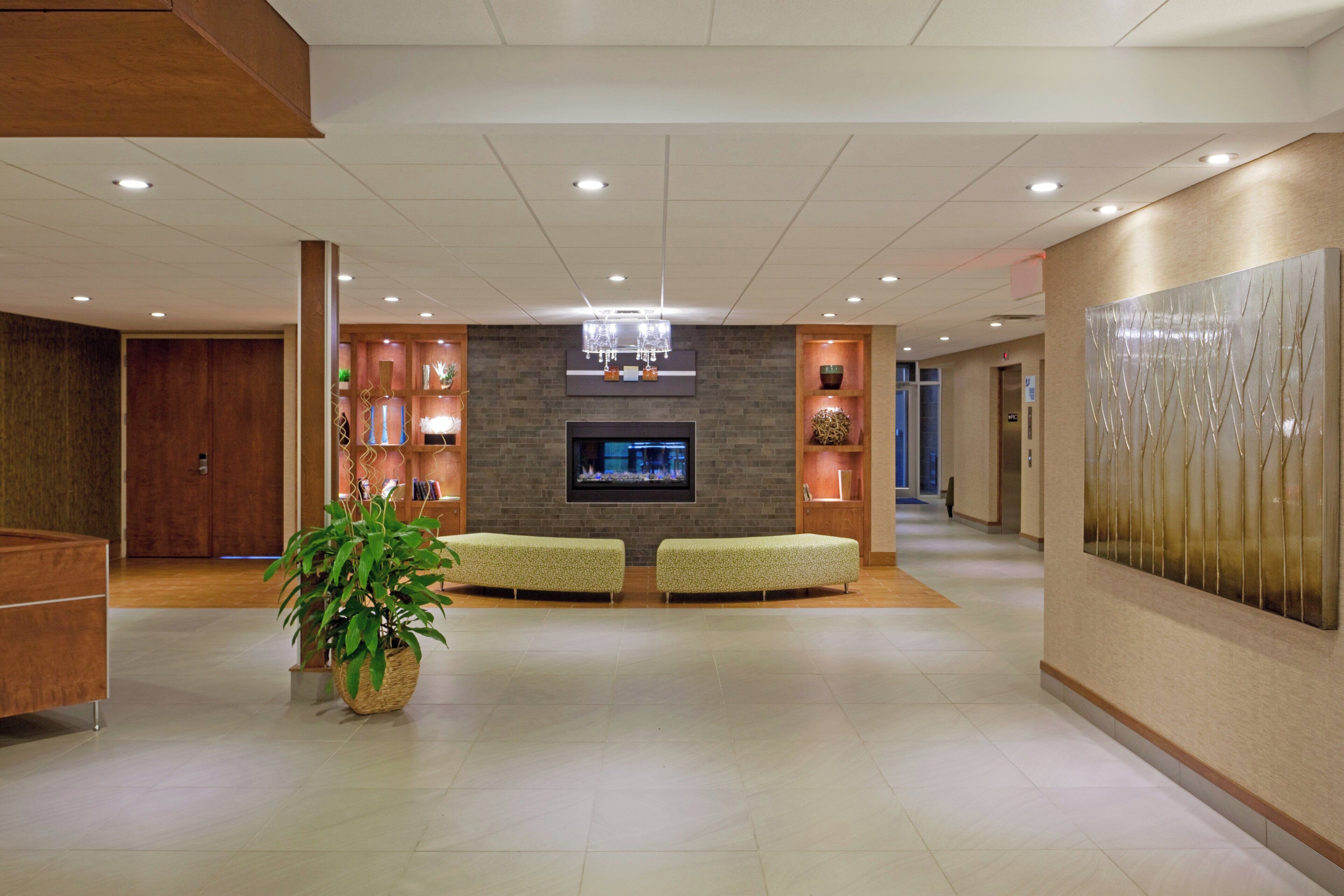 A spacious lobby with a flat-screen TV, two green cushioned seating areas, a large potted plant, and a decorative wall panel.