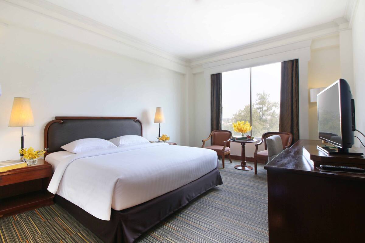 A hotel room with a large bed, two bedside lamps, a desk with a television, and a view of trees through the window.
