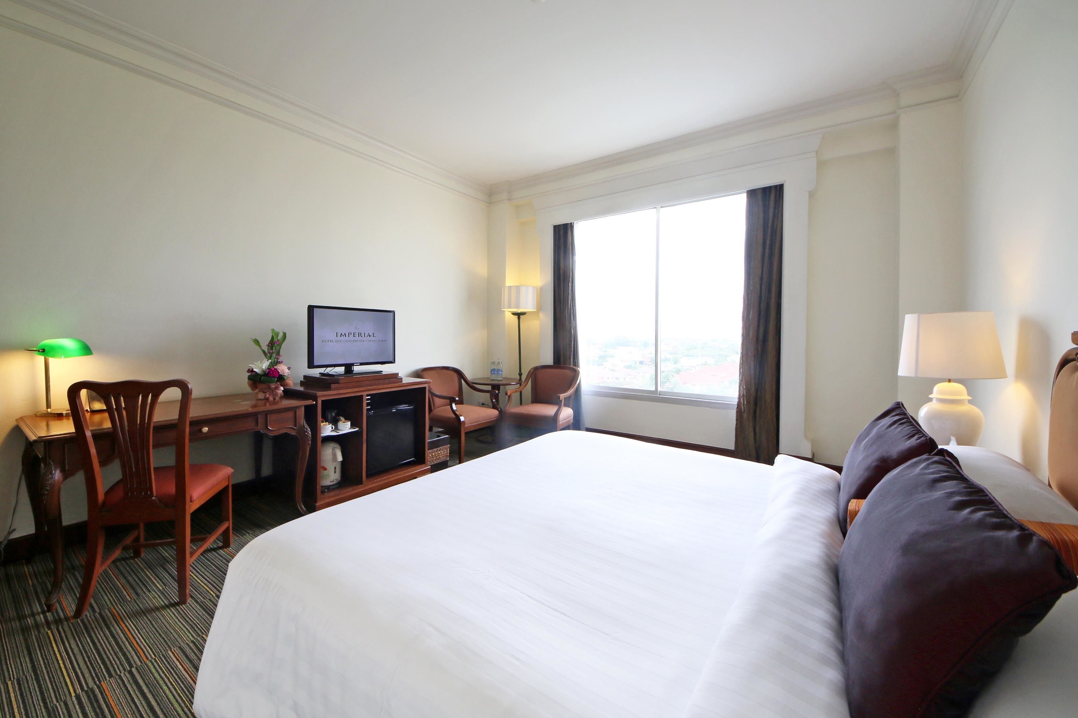 A hotel room with a large bed, a desk with a computer, a chair, and a window with curtains.