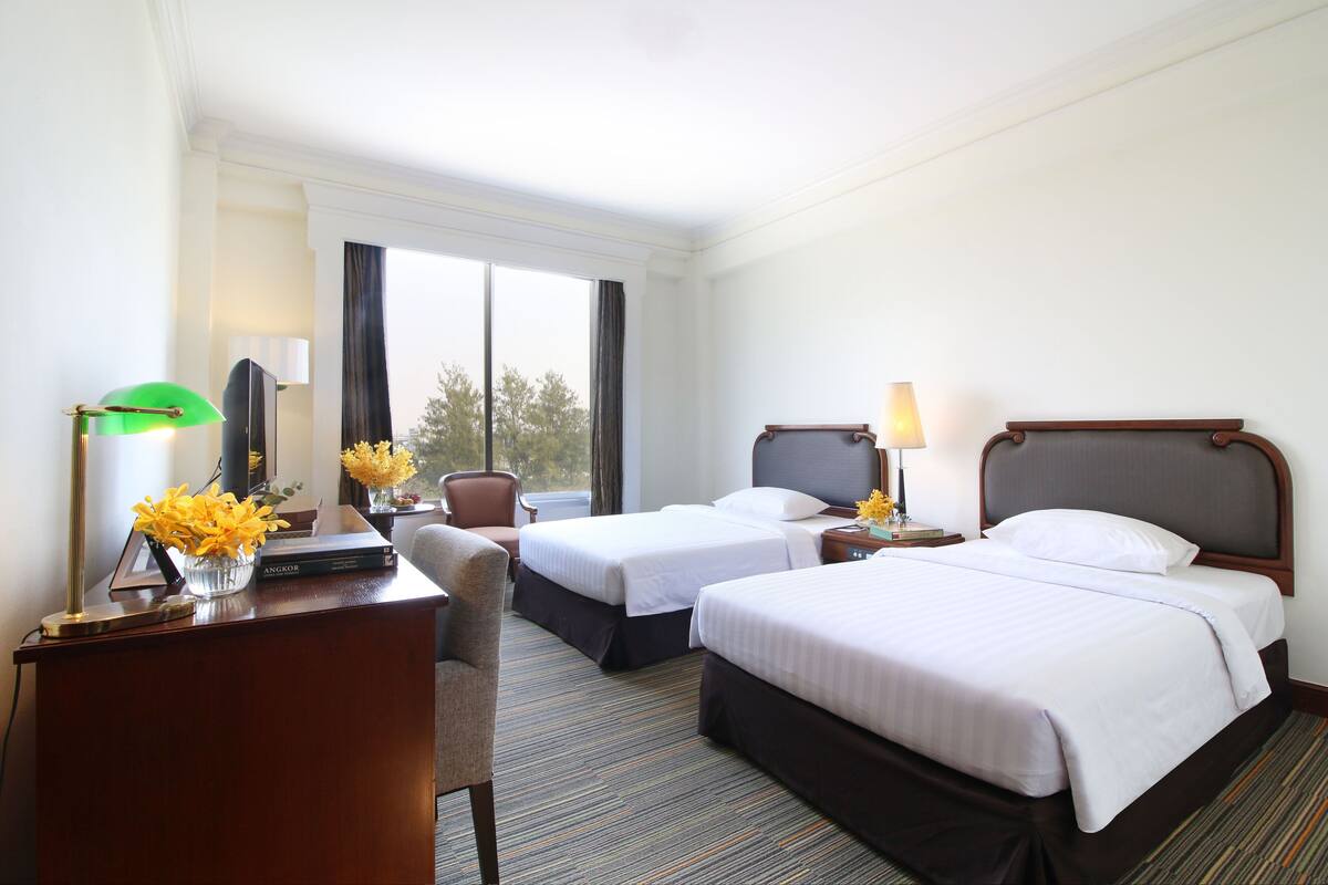 A hotel room with two beds, a desk with a lamp, a chair, and a window with curtains.