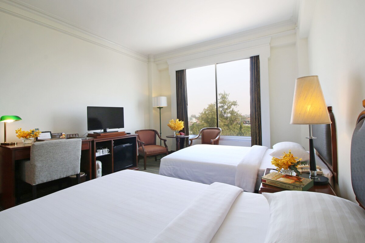 A hotel room with two beds, a desk with a computer, a TV, a lamp, and a window with a view.