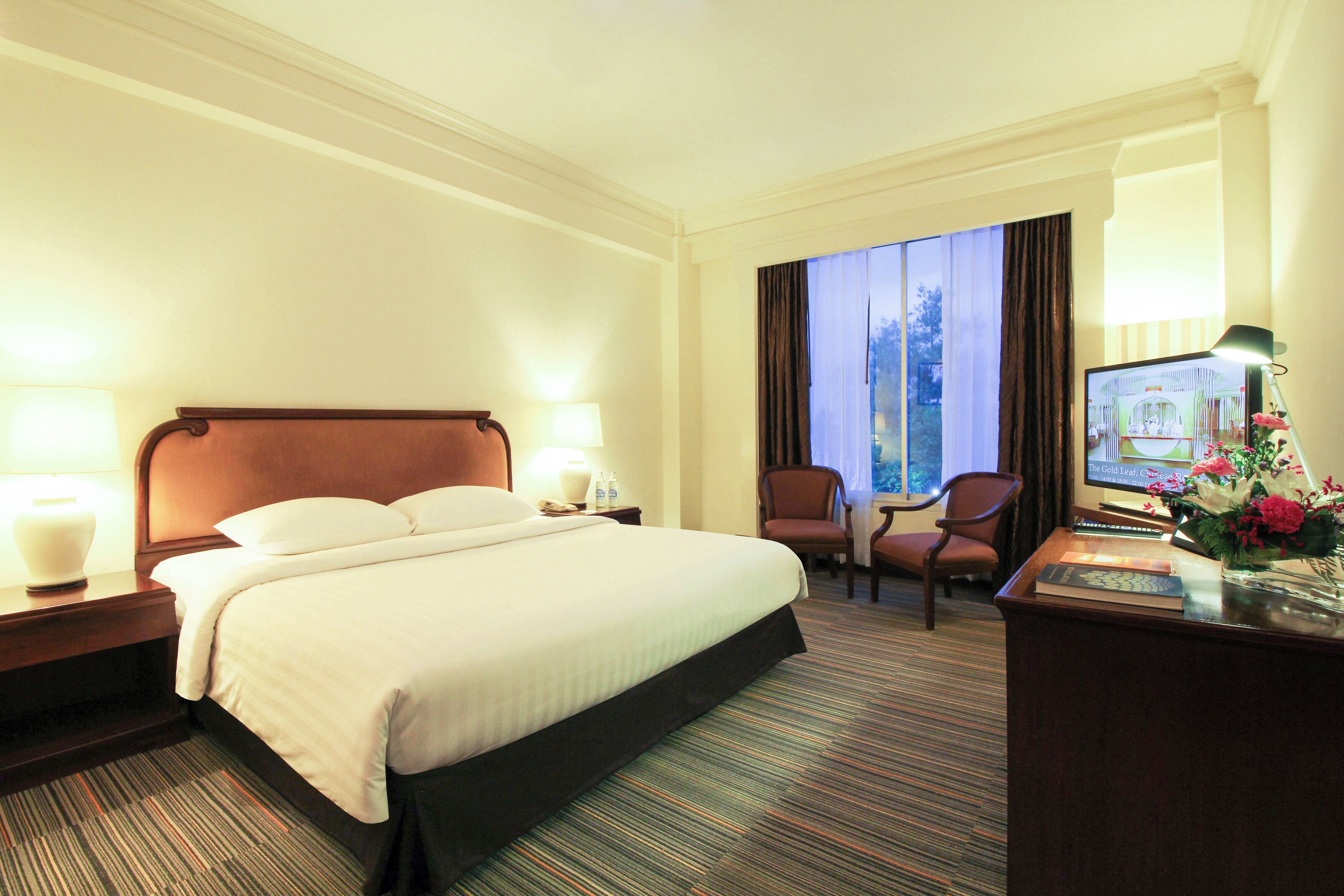 A hotel room with a large bed, a desk with a computer, a chair, and a window with curtains.
