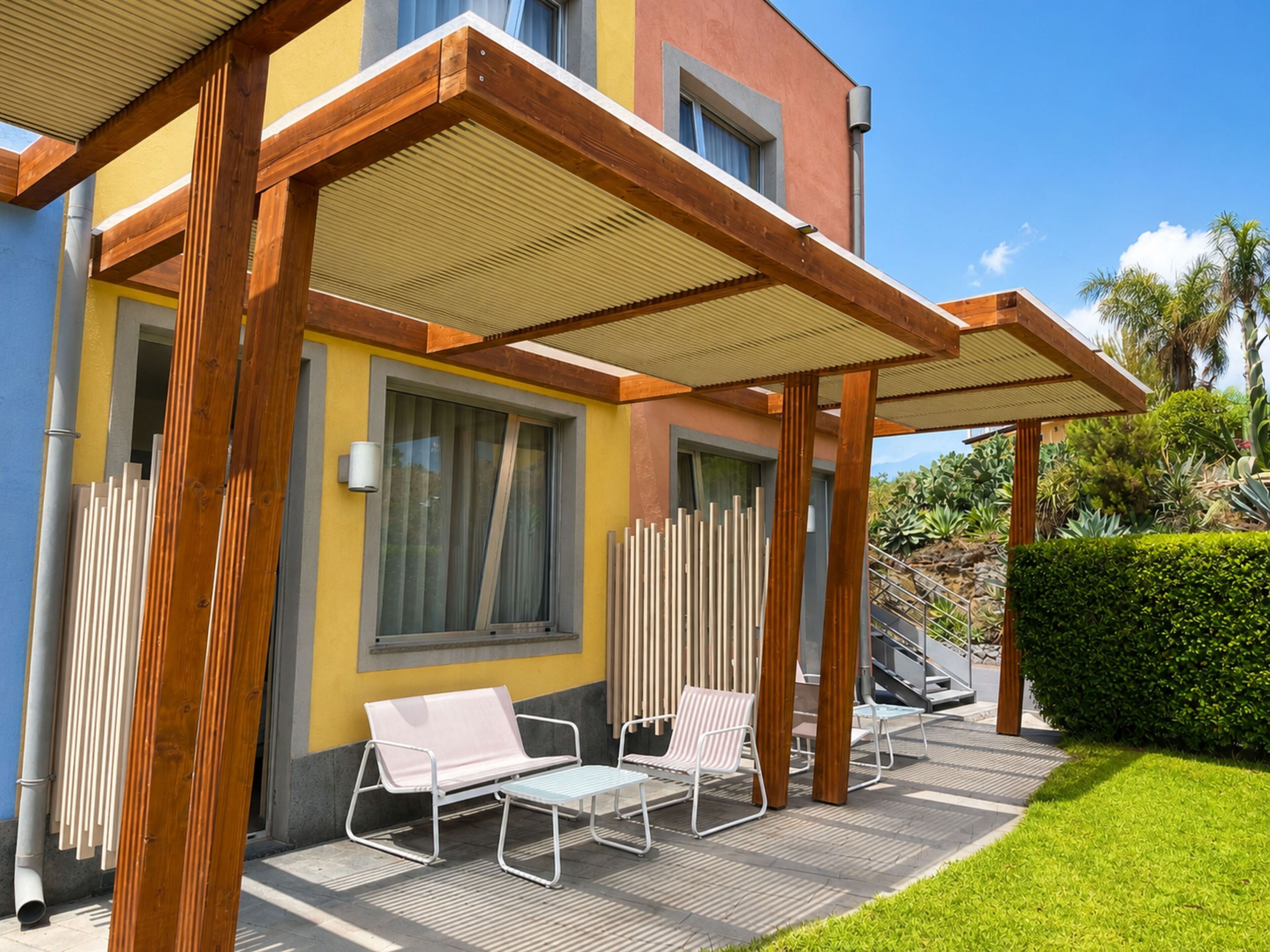 A modern outdoor seating area with a pergola, two lounge chairs, and a staircase leading to a building with a yellow facade.