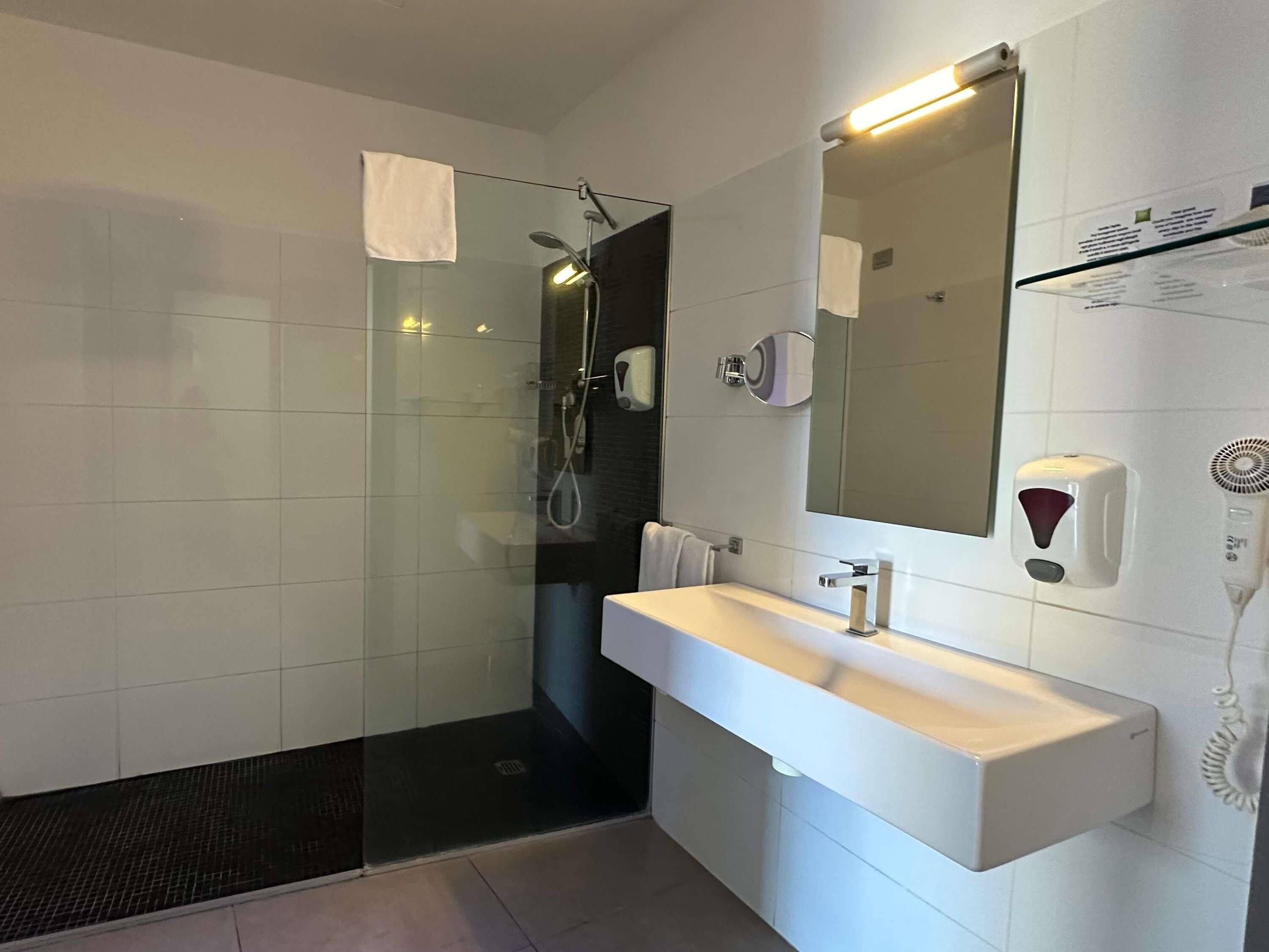 A modern bathroom with a glass shower enclosure, a white sink, a mirror, and a hand dryer.