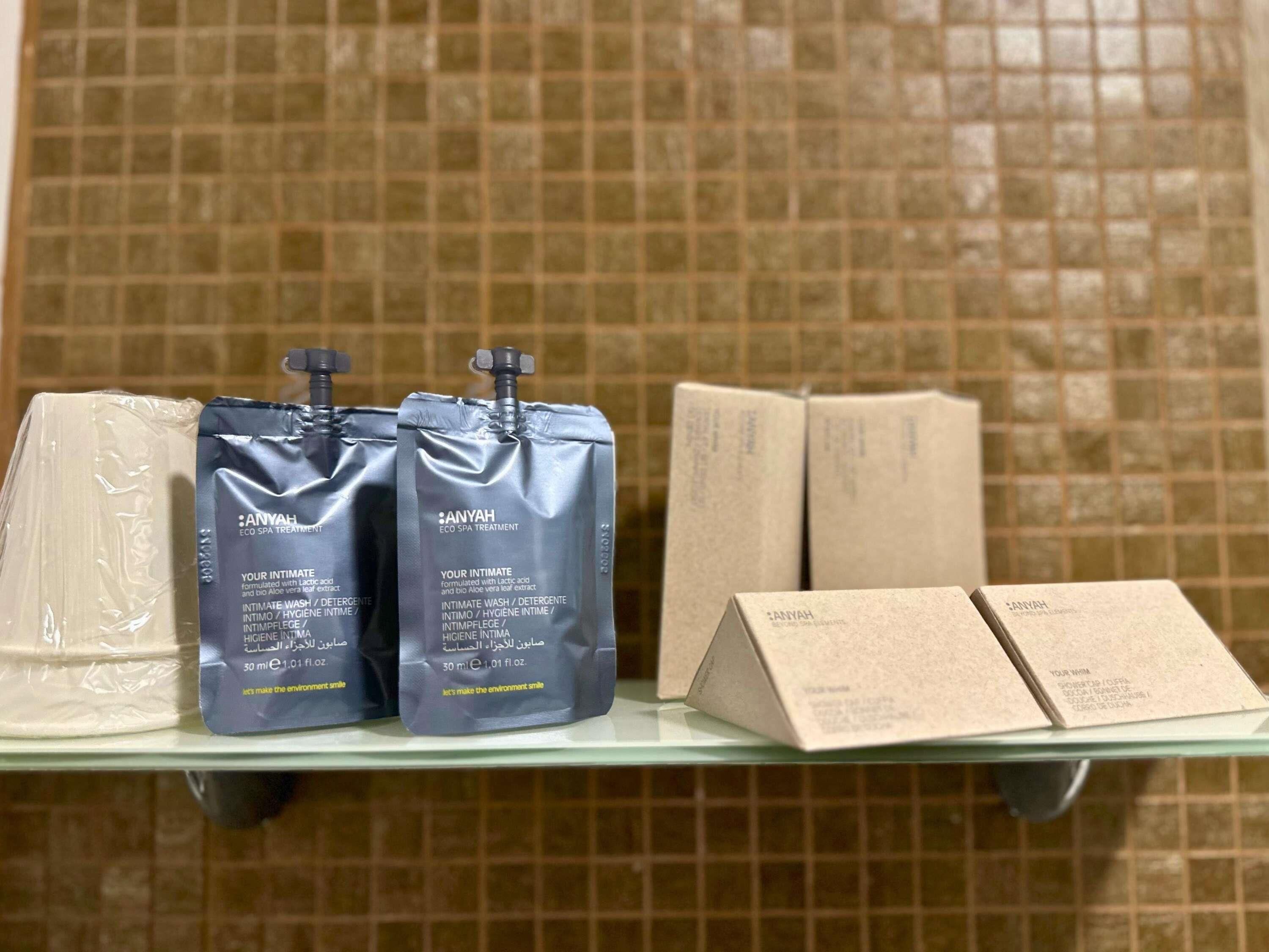 Casa de banho com parede revestida em azulejos, prateleira de vidro e vários produtos de higiene pessoal, incluindo duas garrafas de Anyah Eco Spa Treatment, uma barra de sabonete e duas caixas.