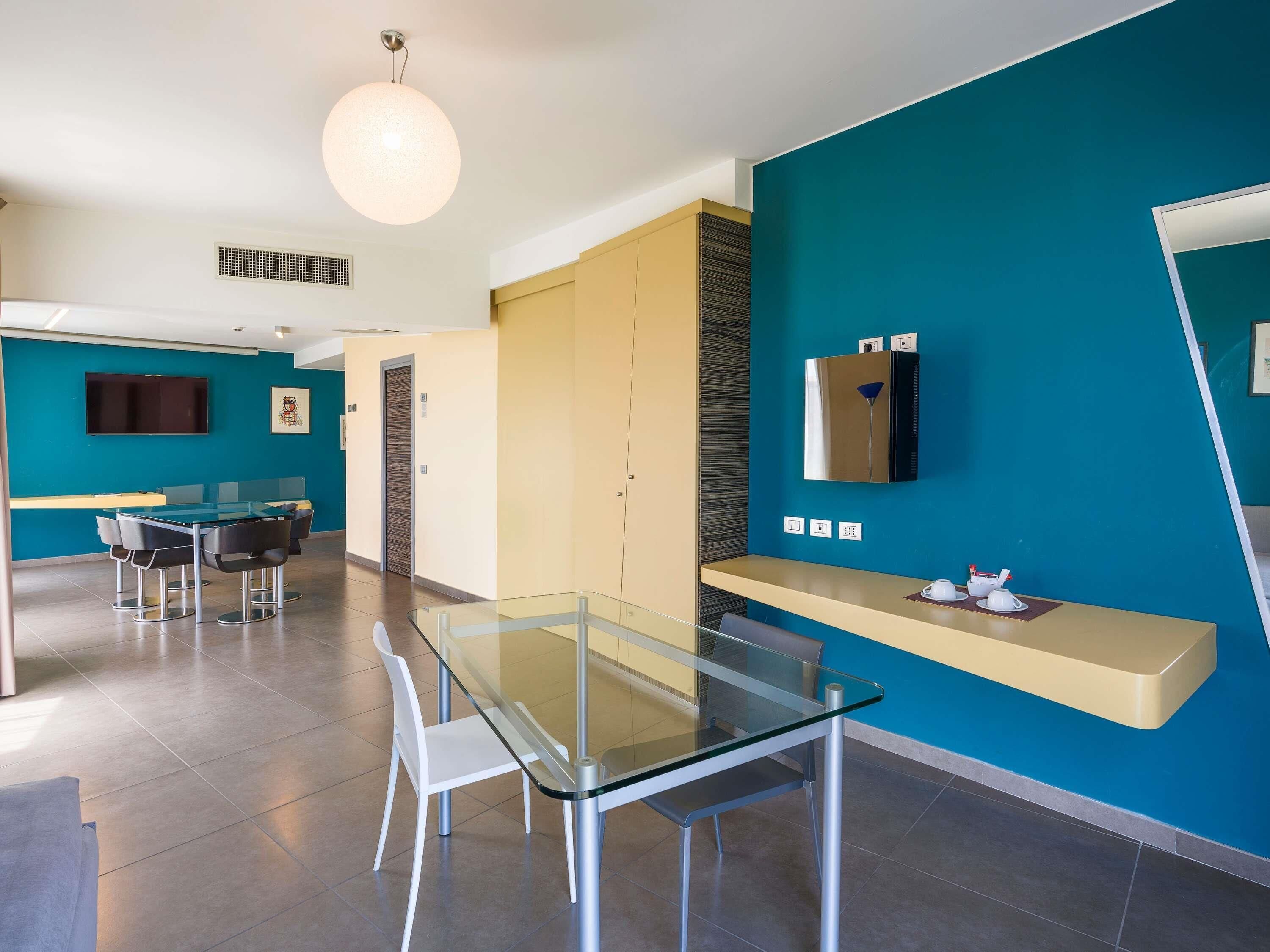 A modern hotel room with a glass dining table, white chairs, a flat-screen TV, and a minibar.