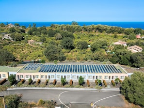 A large building with a rooftop solar panel array, surrounded by greenery and a clear view of the sea.