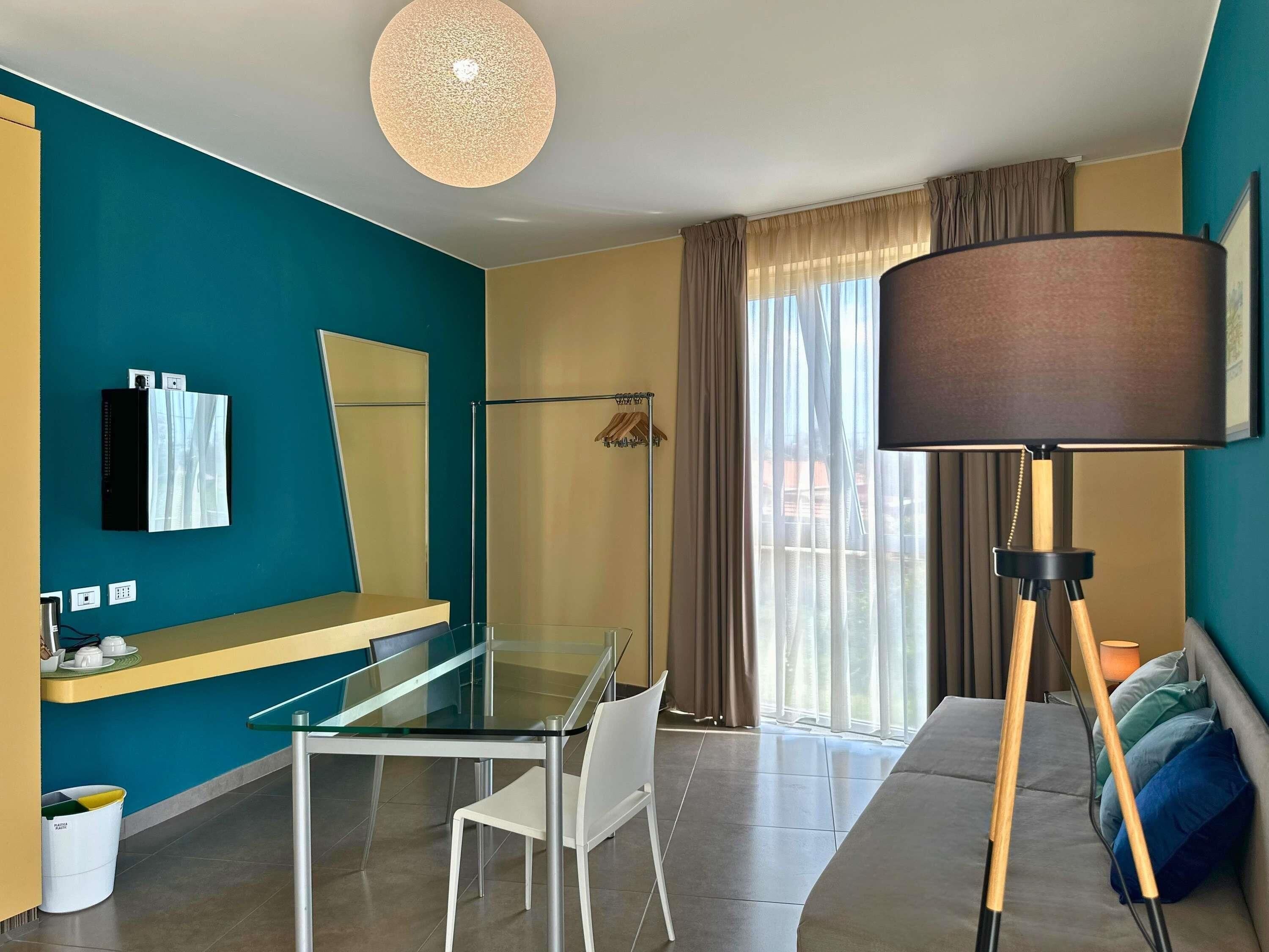 A modern hotel room with a glass table, white chairs, a sofa, a large floor lamp, and a view of the outdoors through a window with curtains.