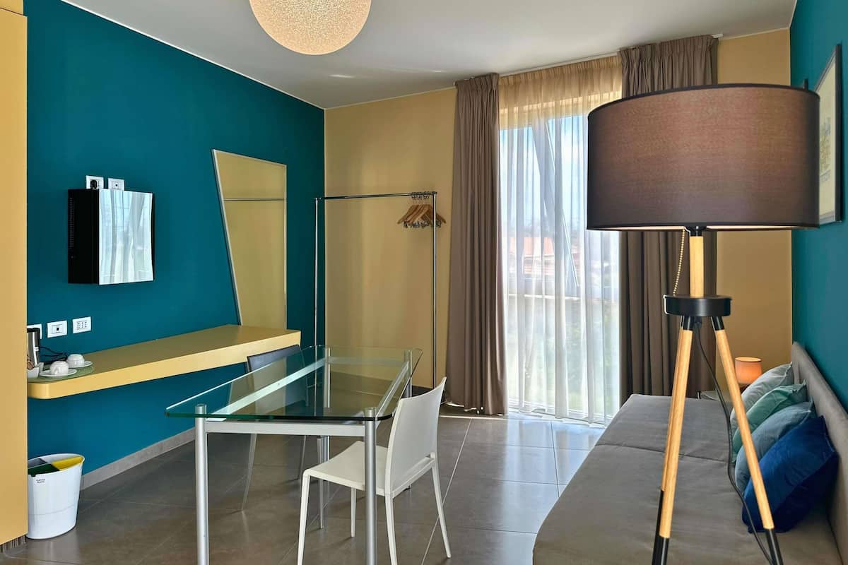 A modern hotel room with a glass table, white chairs, a sofa, a large floor lamp, and a view of the outdoors through a window with curtains.