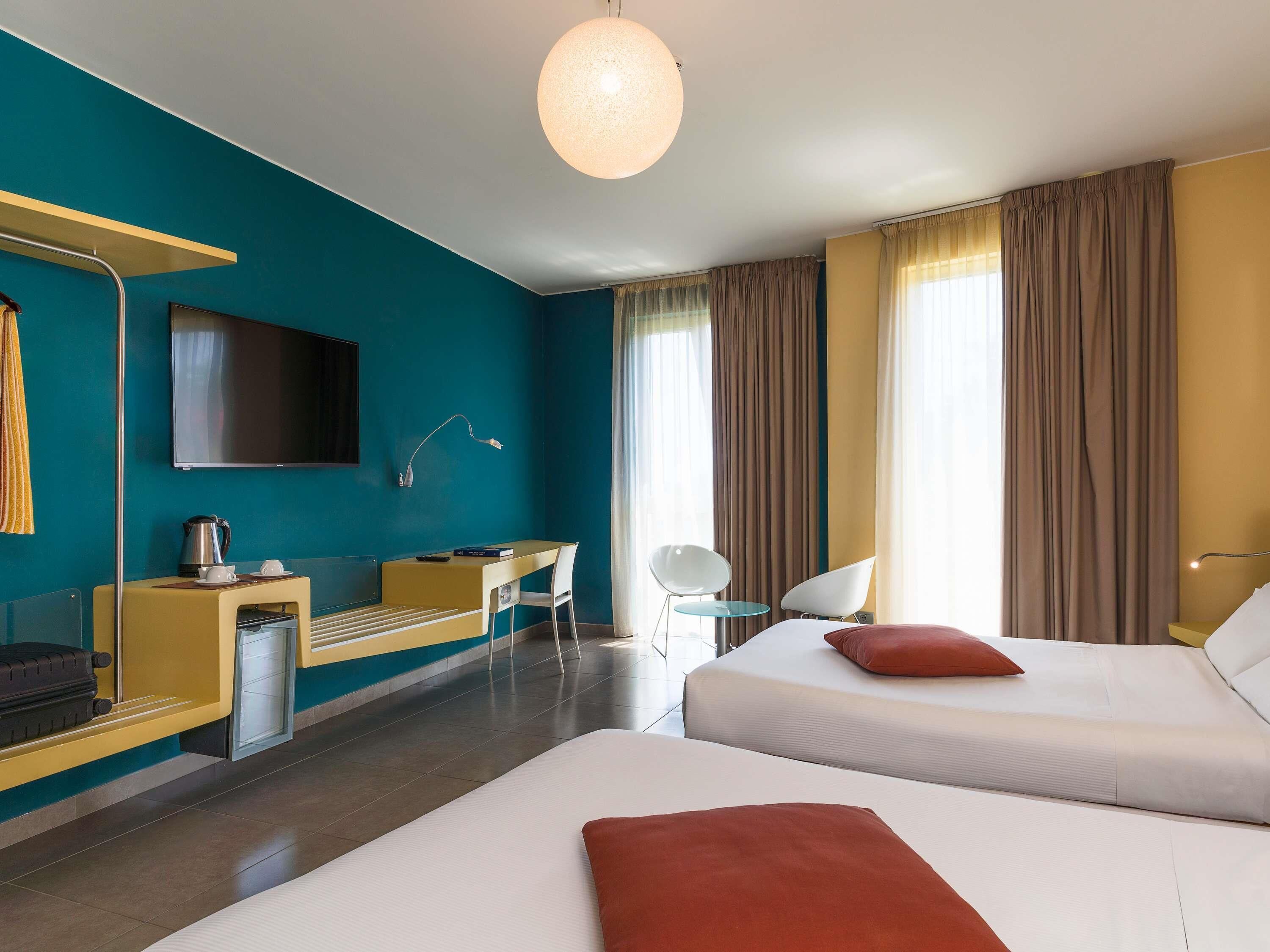 Habitación de hotel con cama, escritorio, silla, televisor y una ventana grande con cortinas.