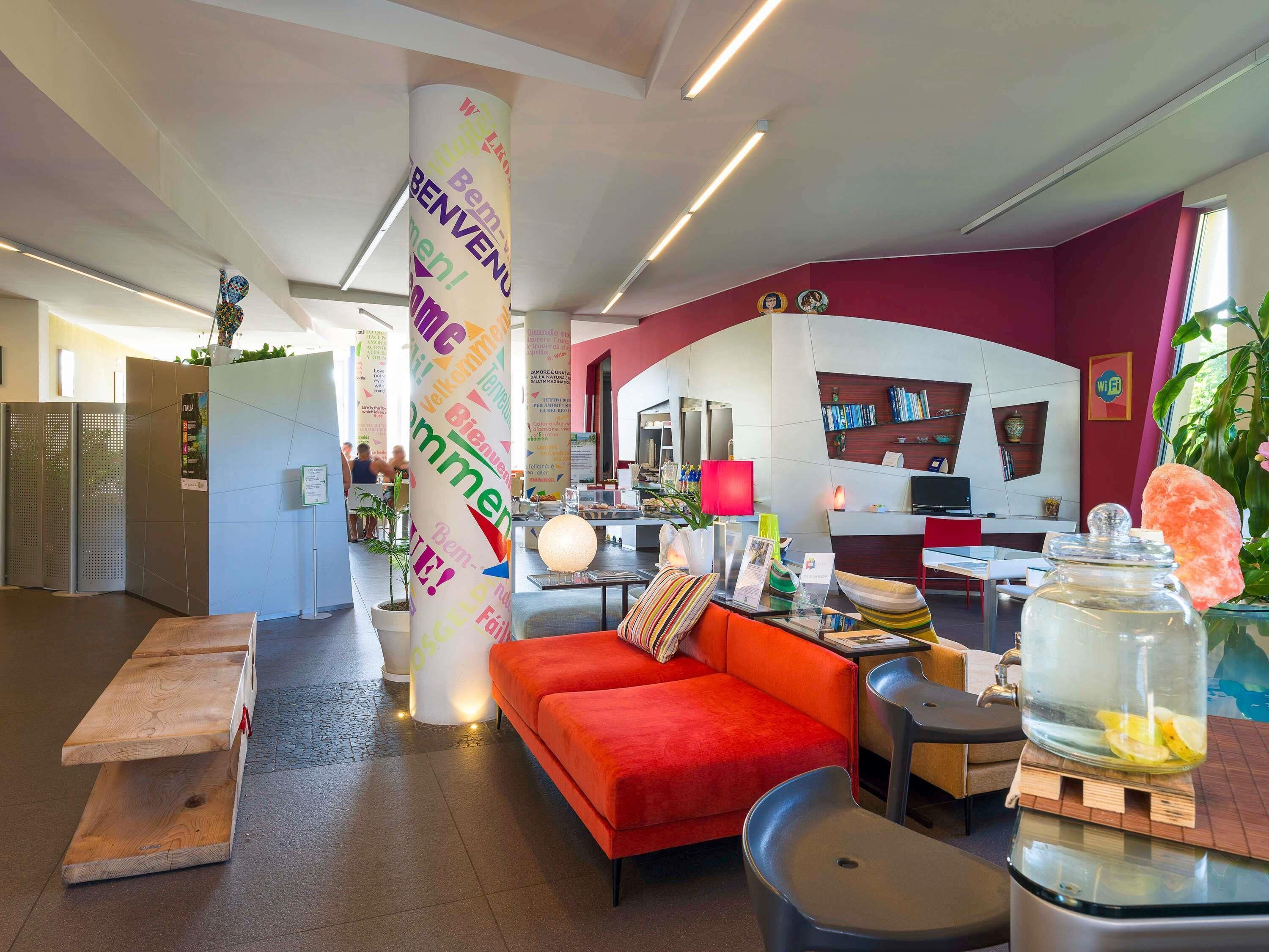 A modern hotel lobby with a red sofa, a wooden bench, a glass table with a jar of lemons, and a colorful pillar with 'Welcome' text.